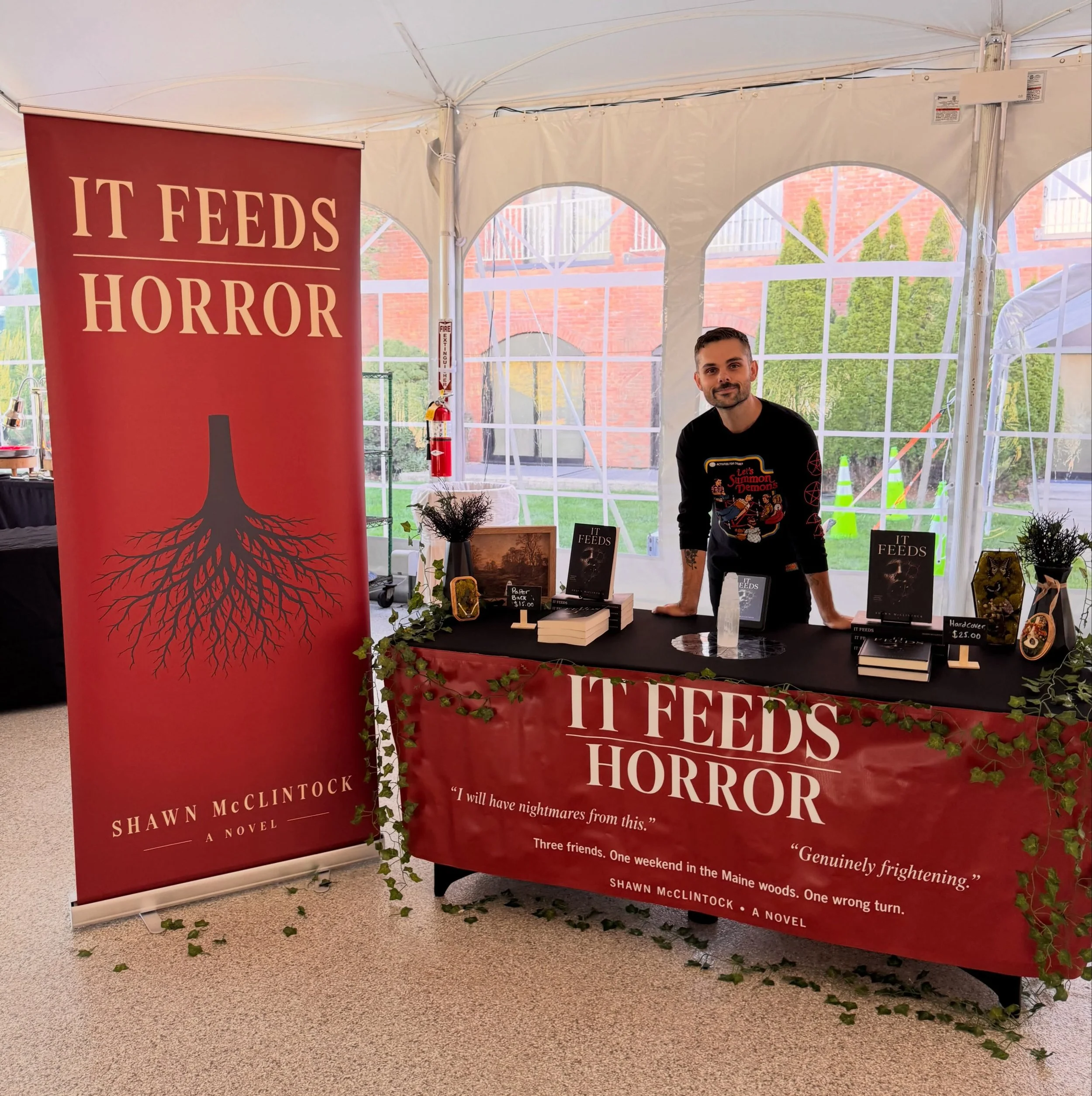 Author Shawn McClintock at a book display for his horror novel It Feeds Horror, surrounded by promotional banners and copies of the book.