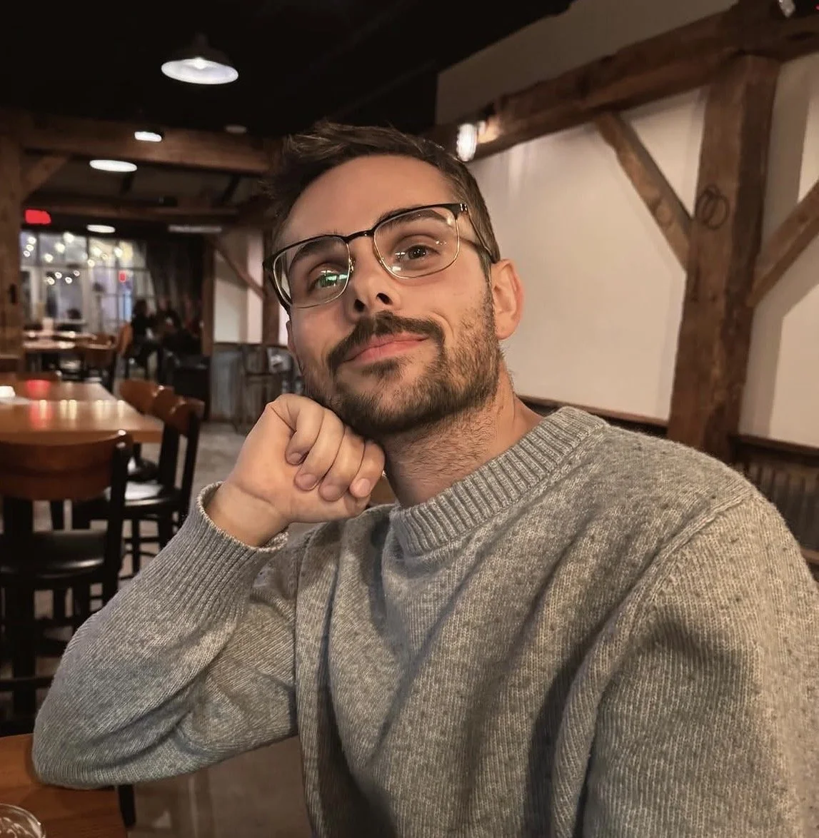 Man wearing glasses and a gray sweater smiles for a selfie in a warmly lit restaurant.