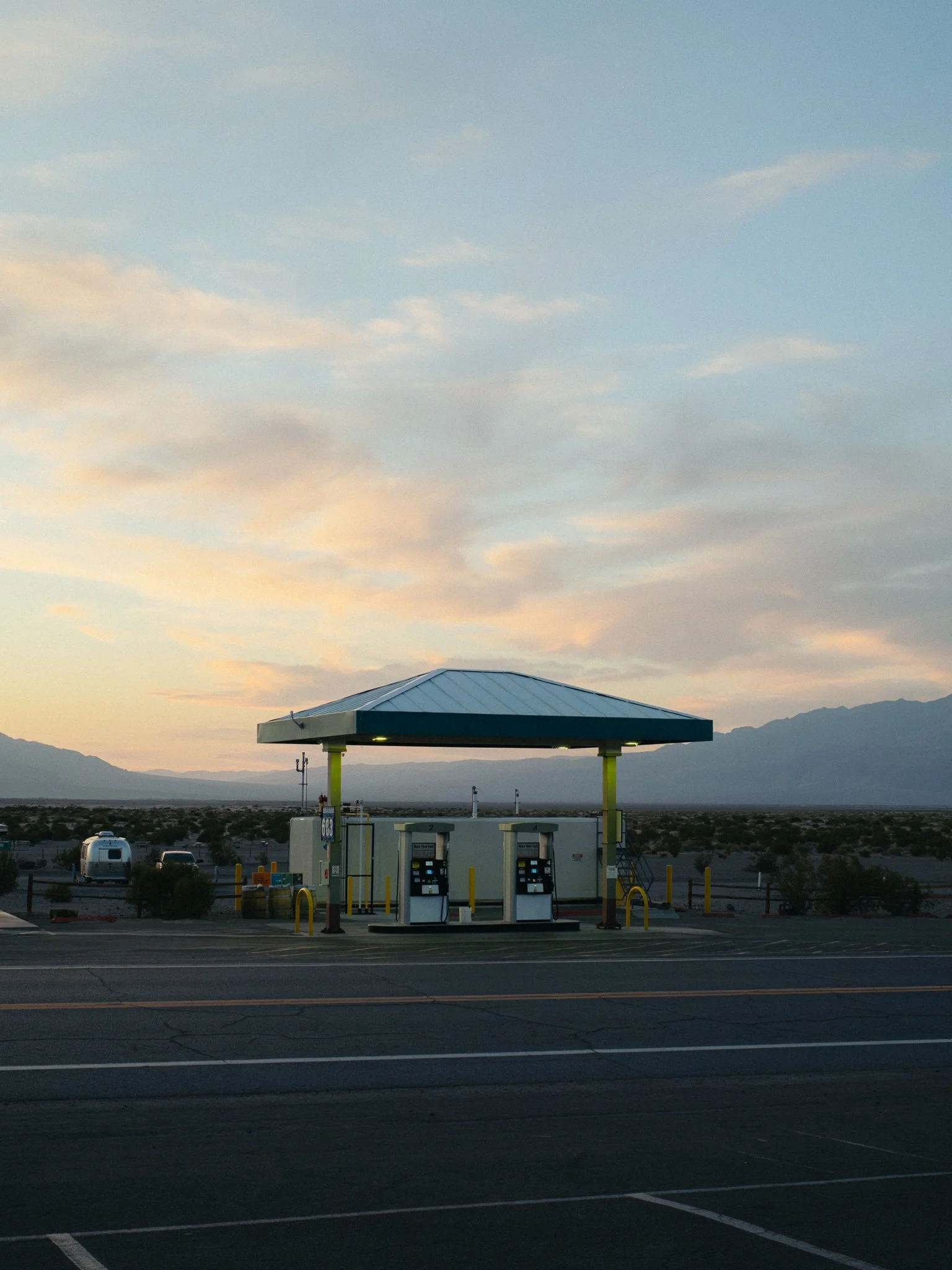 Gas station pumps beneath a canopy at sunset, with desert landscape and mountains in the background