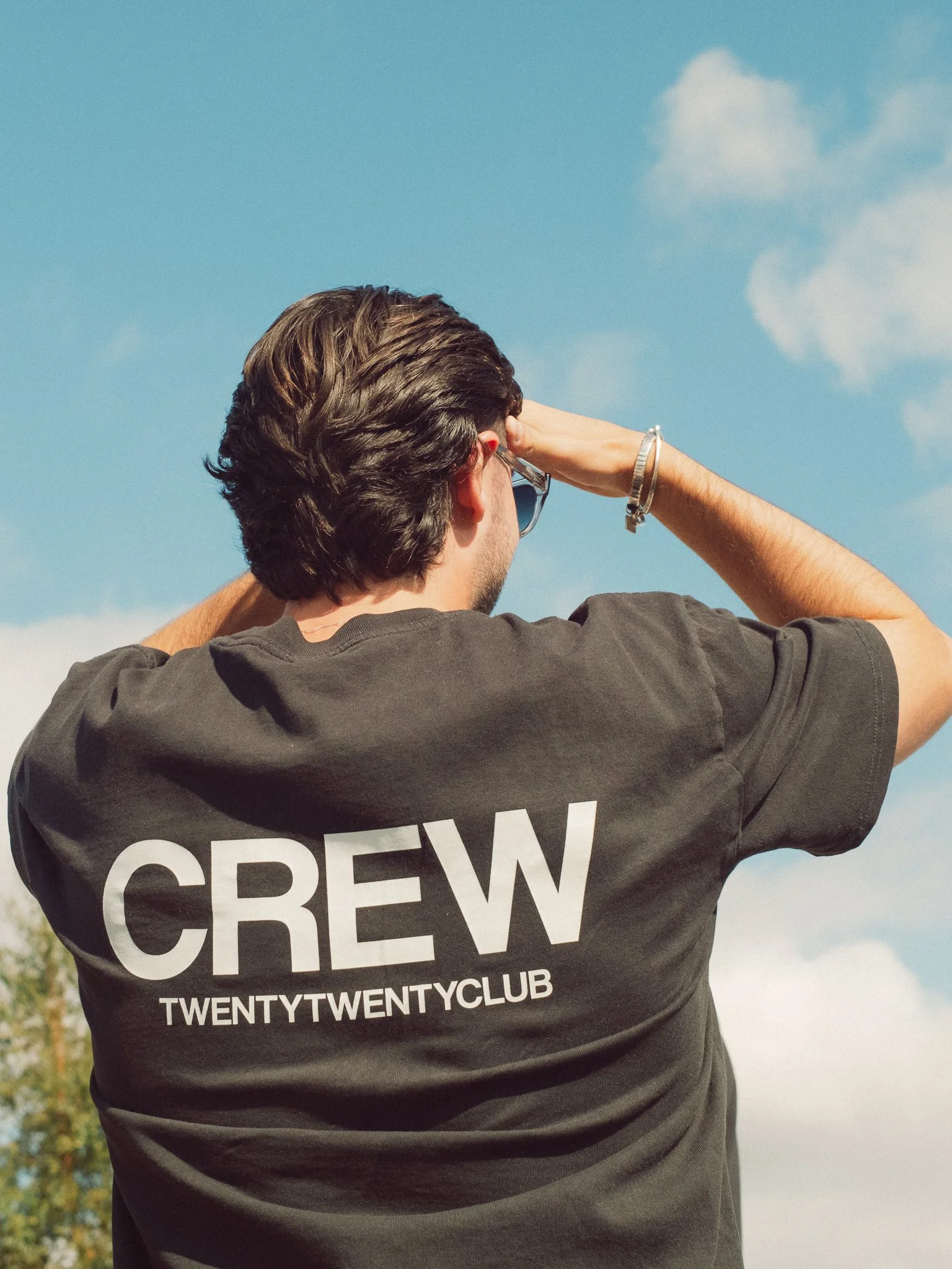 Person wearing a black CREW TwentyTwenty Club T-shirt and sunglasses under a blue sky