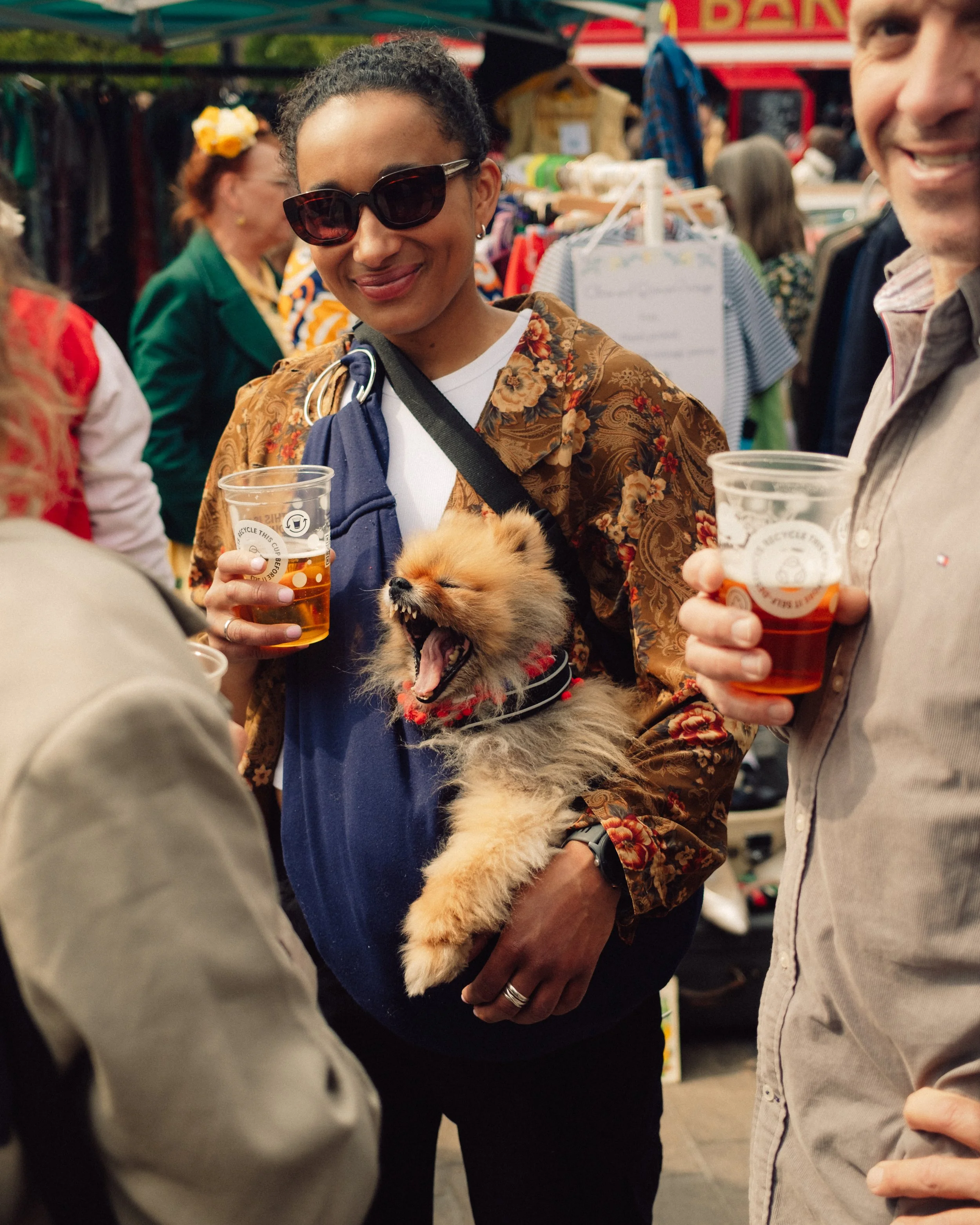 Woman wearing sunglasses holds a small dog at a crowded outdoor market while friends hold plastic cups of drinks.