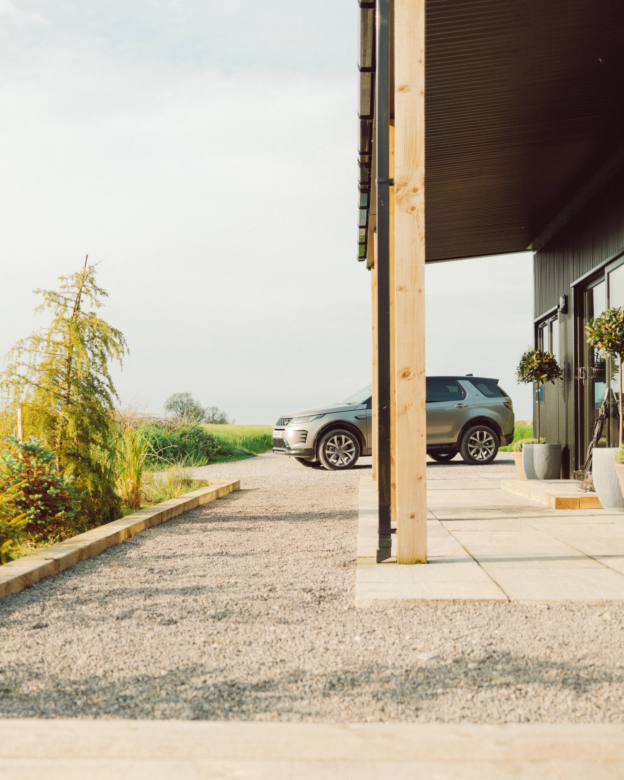 Modern home exterior with wood posts, landscaped garden, gravel driveway, and parked silver SUV