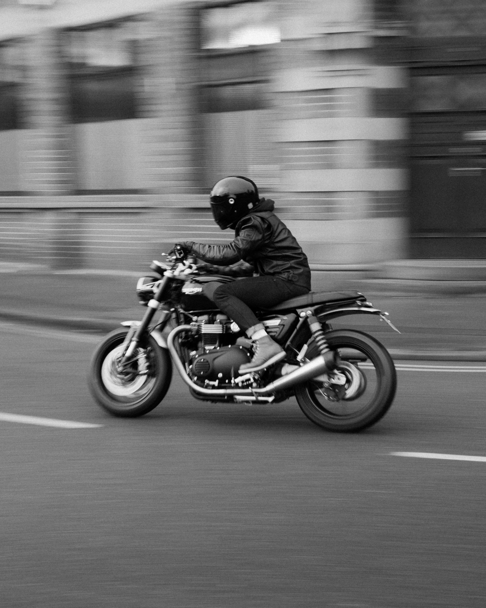 Black-and-white motion-blur photo of a helmeted motorcyclist riding a Triumph motorcycle on a city street