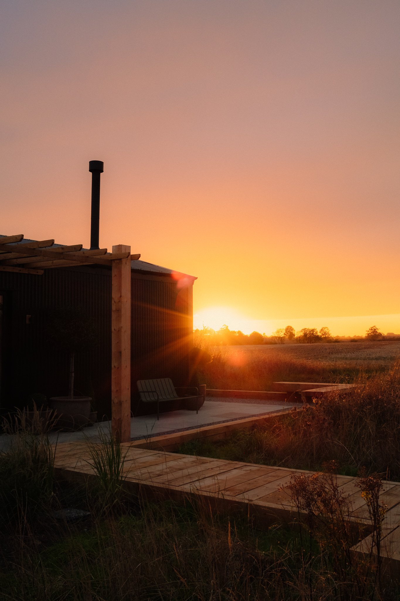 Sunset over a rural field beside a rustic corrugated-metal shed, wooden deck, and outdoor seating.