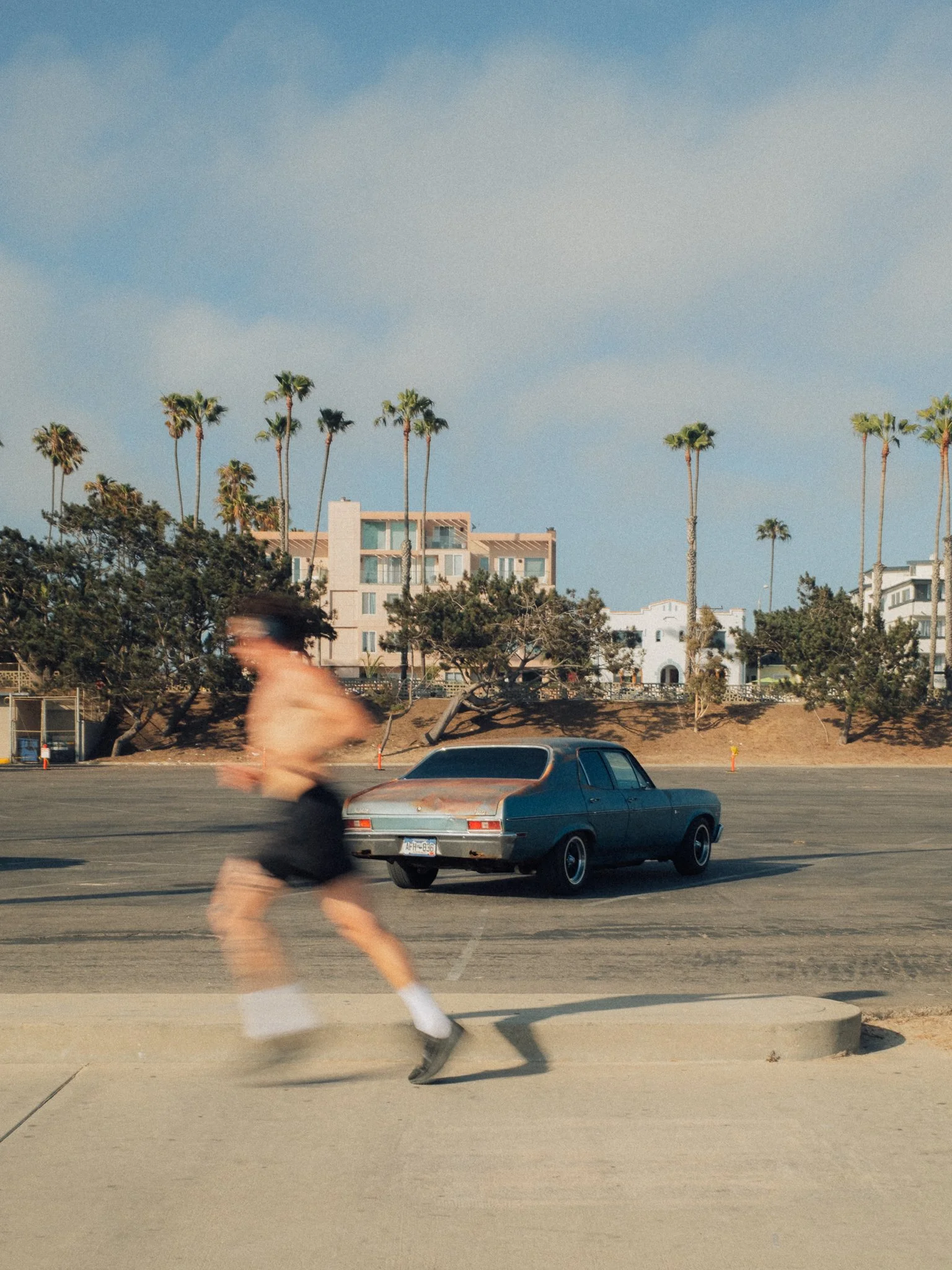 Motion-blurred person walking past a vintage blue car on a sunny Southern California street lined with palm trees and homes.