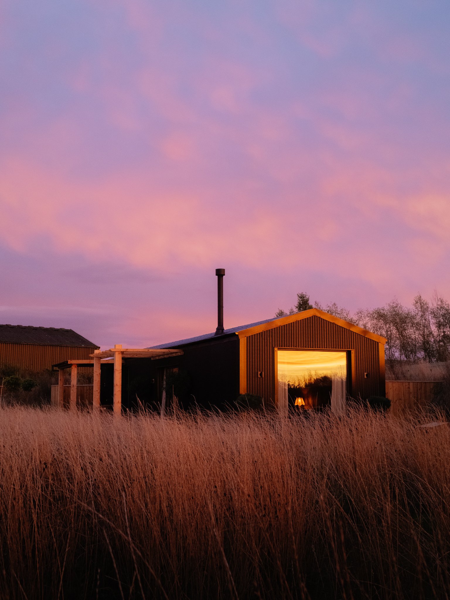Modern black cabin with a chimney and glowing window amid tall grasses at pink and purple sunset