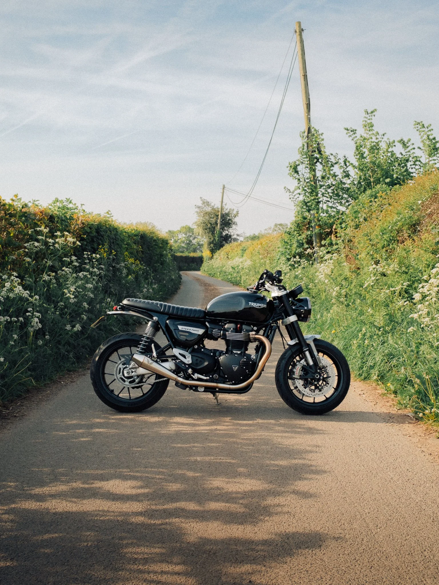 Black Triumph Speed Twin motorcycle parked on a rural lane surrounded by lush hedges and wildflowers