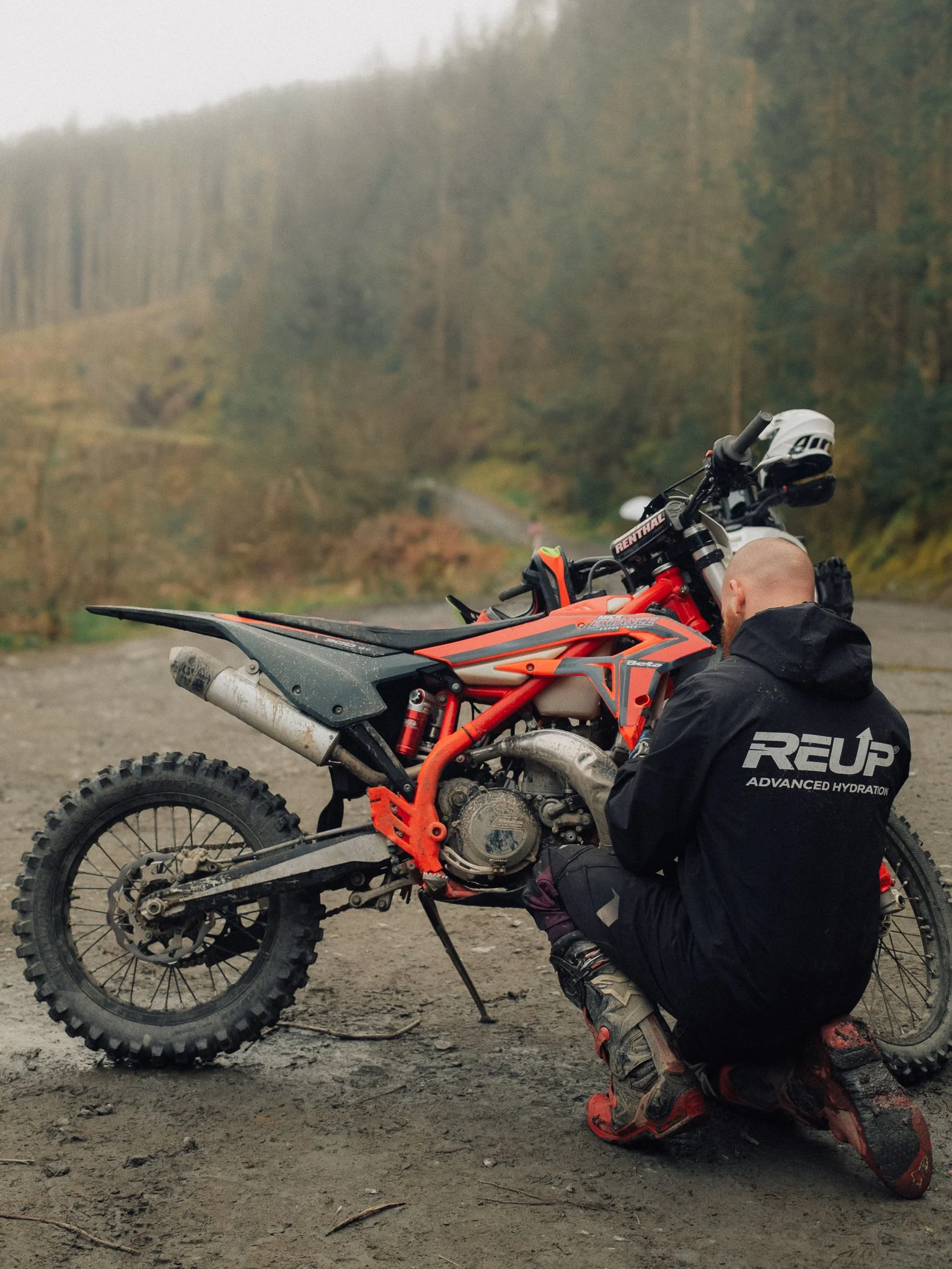 Off-road rider kneeling beside an orange dirt bike on a muddy forest trail