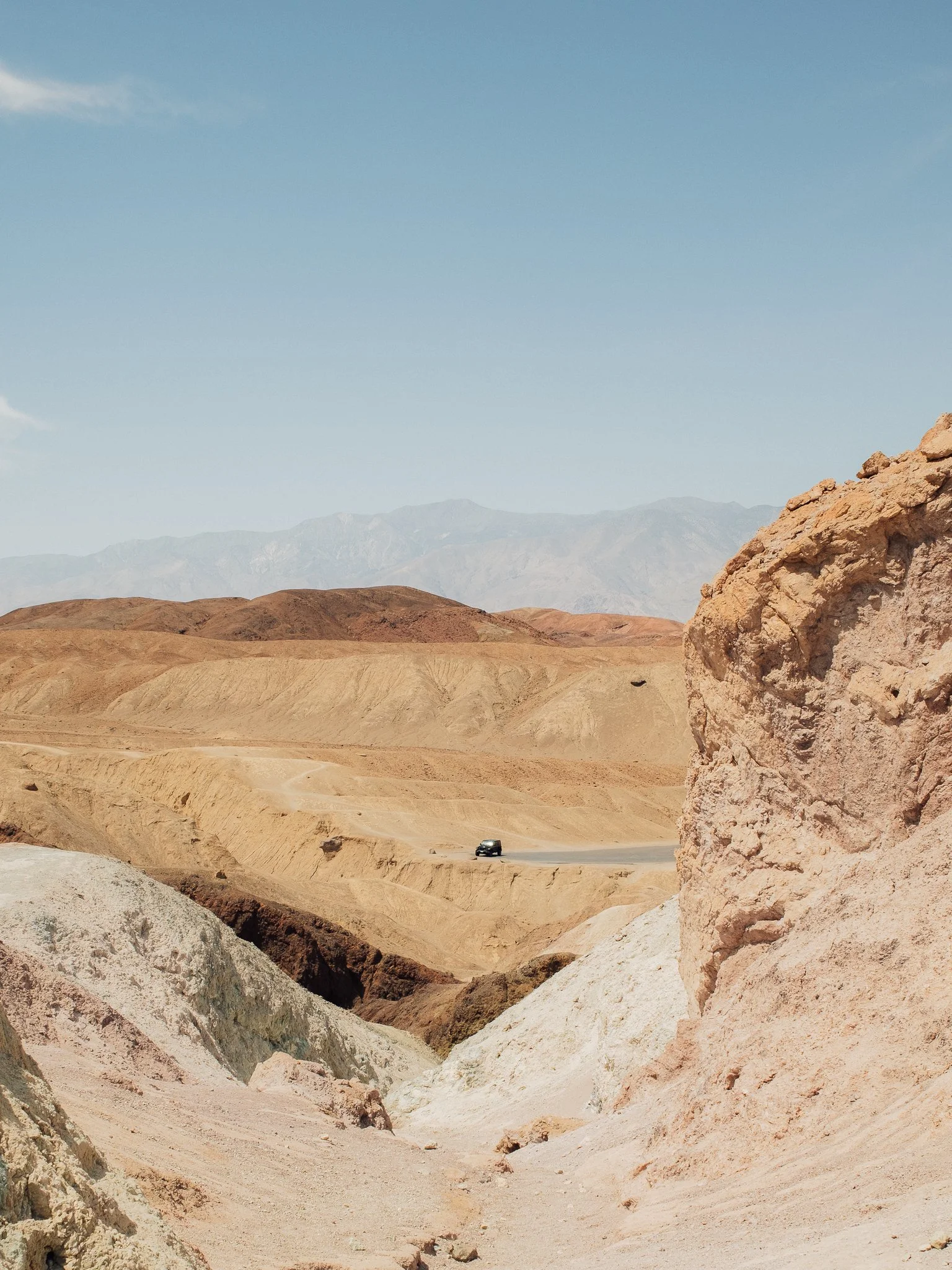Desolate desert landscape with rugged rock formations, winding road, vehicle, and distant mountain range under a clear blue sky