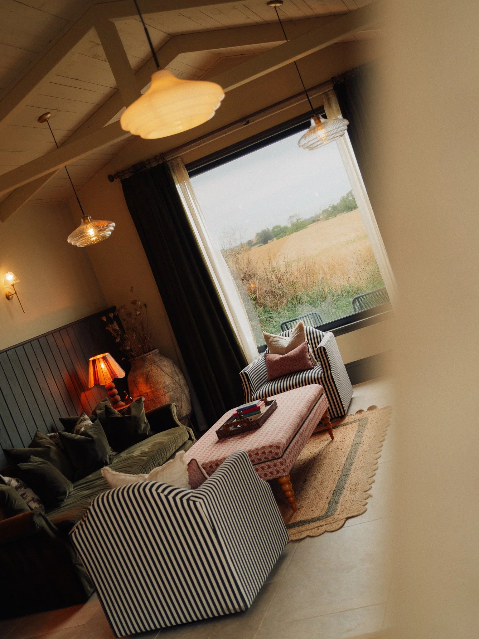 Cozy living room with striped armchairs, sofa, pendant lights, and a large window overlooking a golden field