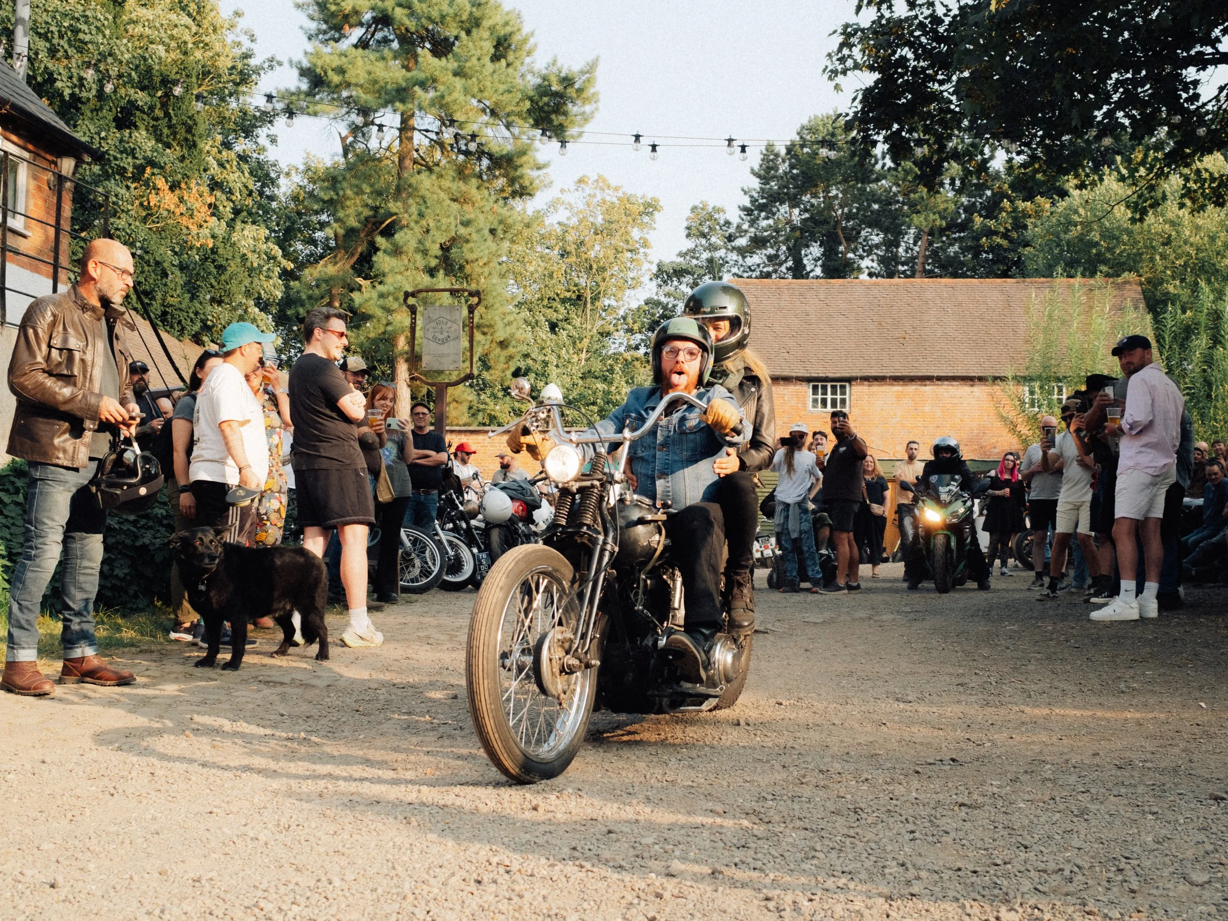 Motorcyclist riding a vintage motorcycle through a crowd at an outdoor gathering with string lights, trees, and brick buildings
