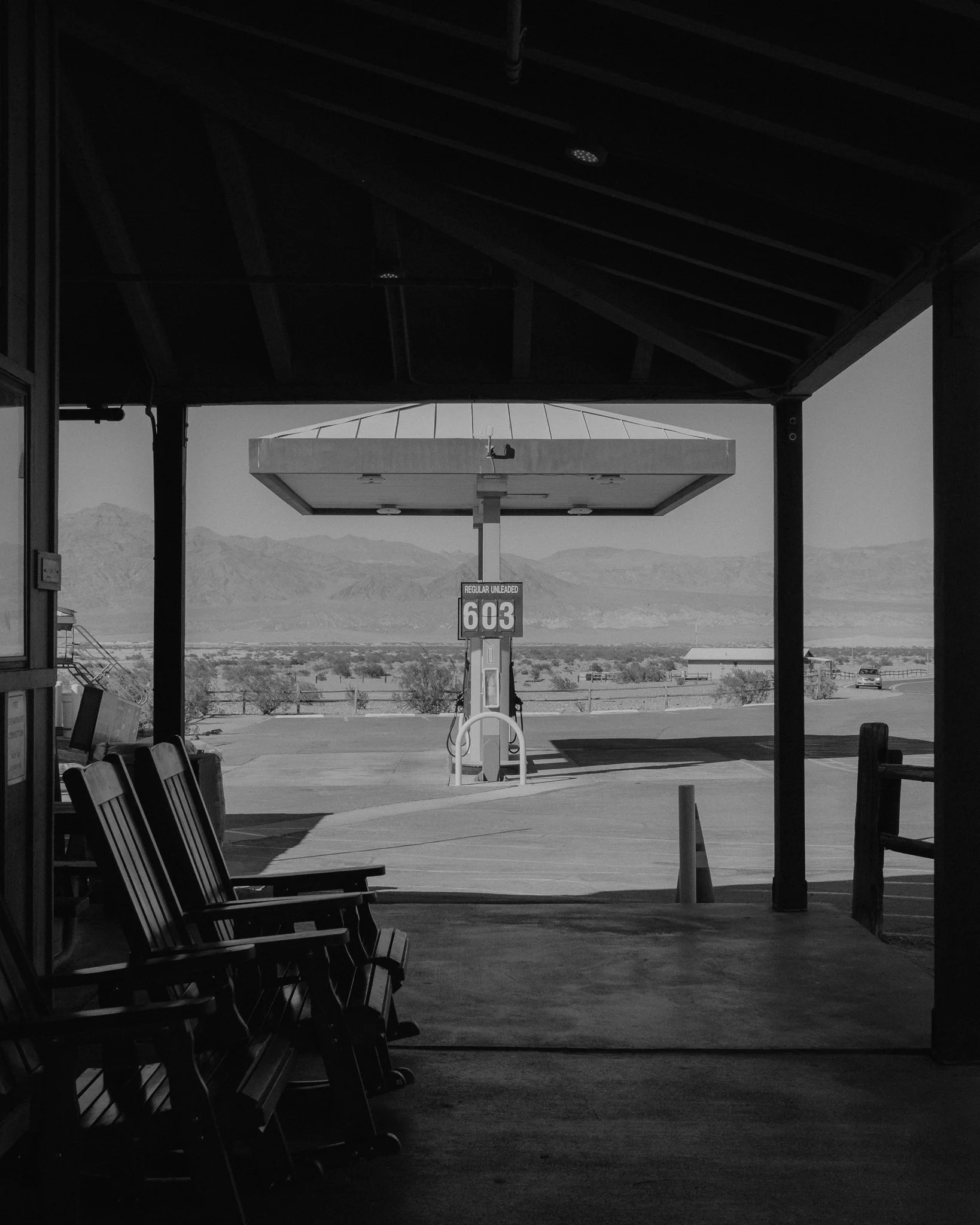 Black-and-white view of a rural gas station pump marked Regular Unleaded 603, with Adirondack chairs and desert mountains in the background
