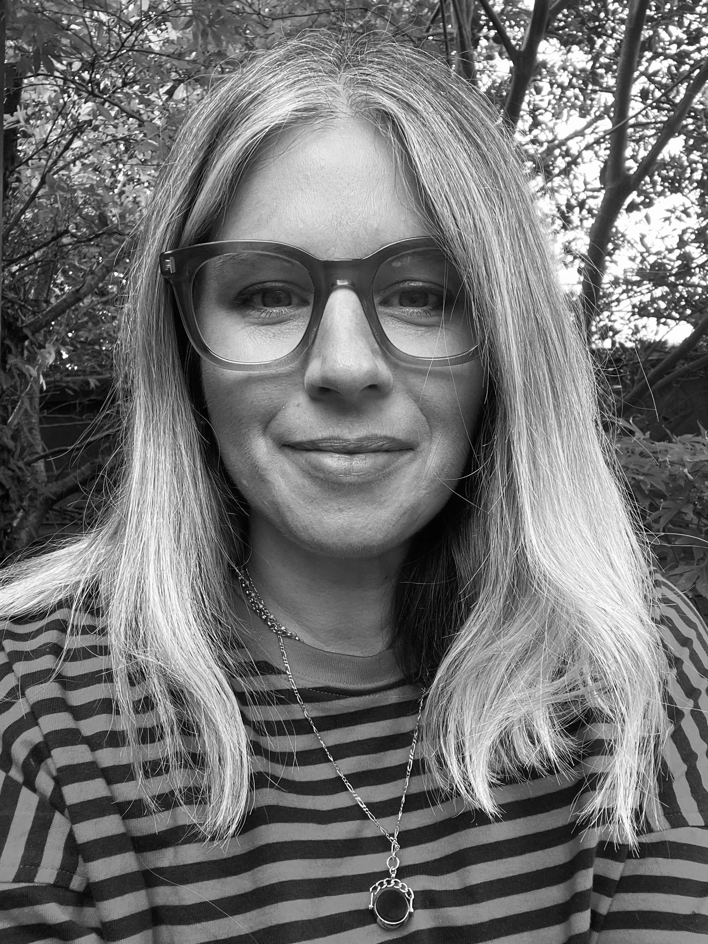 Black-and-white close-up portrait of a woman wearing eyeglasses, a striped top, and a long pendant necklace outdoors among trees.