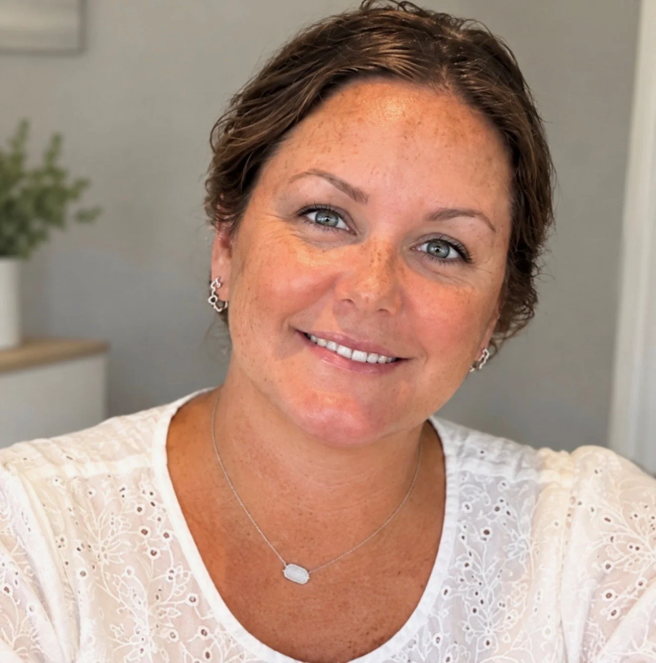 Smiling woman with short brown hair wearing a white embroidered blouse, earrings, and a pendant necklace.