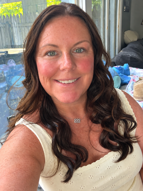 Smiling woman with long wavy brown hair wearing a cream sleeveless top and pendant necklace