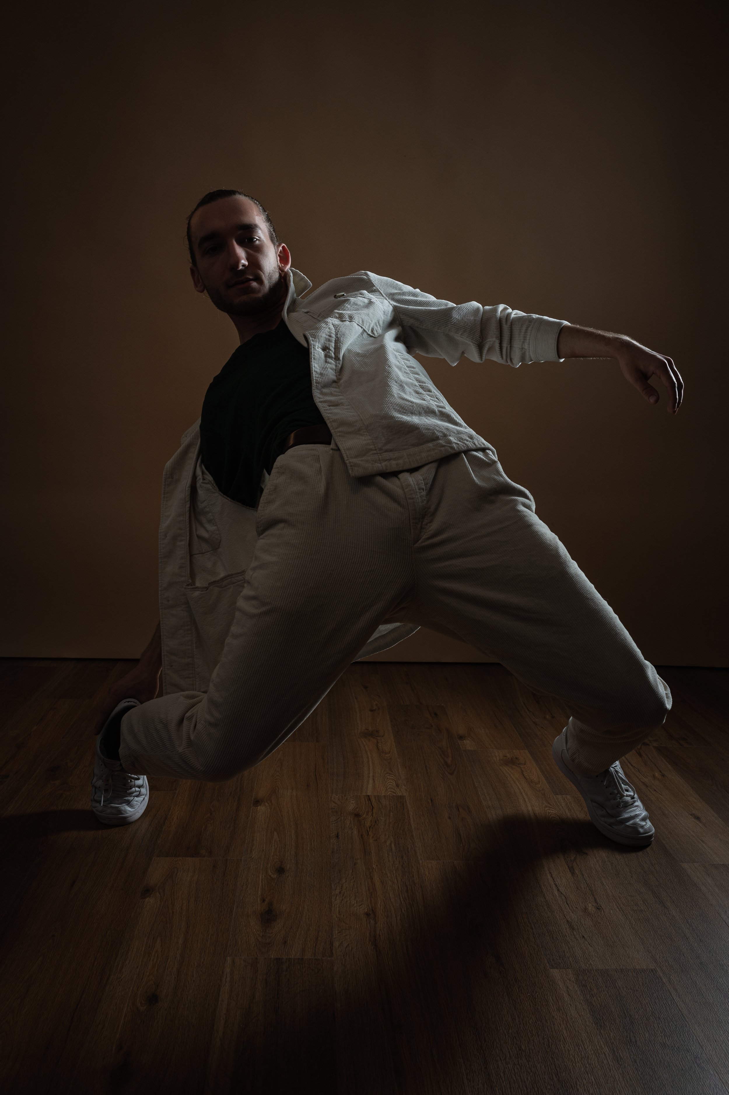 Man in a white outfit crouching with arms extended on a wooden floor against a brown wall