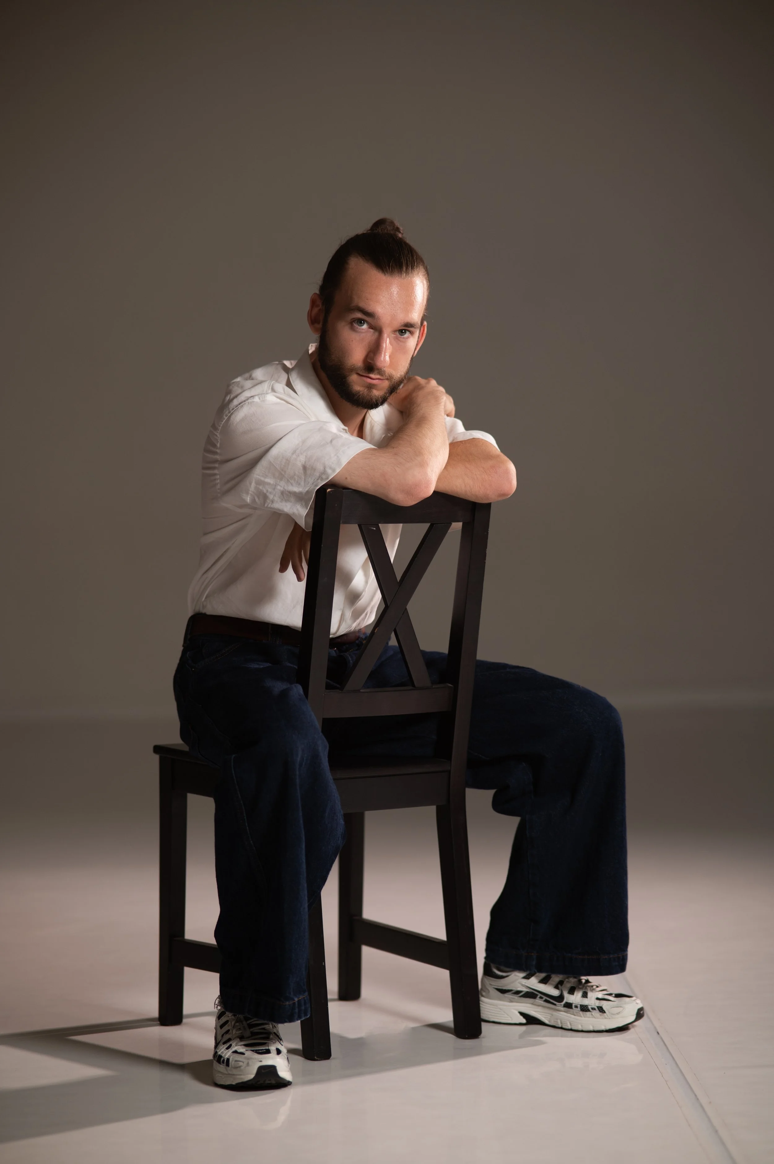 Bearded man with a topknot sitting backward on a wooden chair, wearing a white shirt, jeans, and sneakers.
