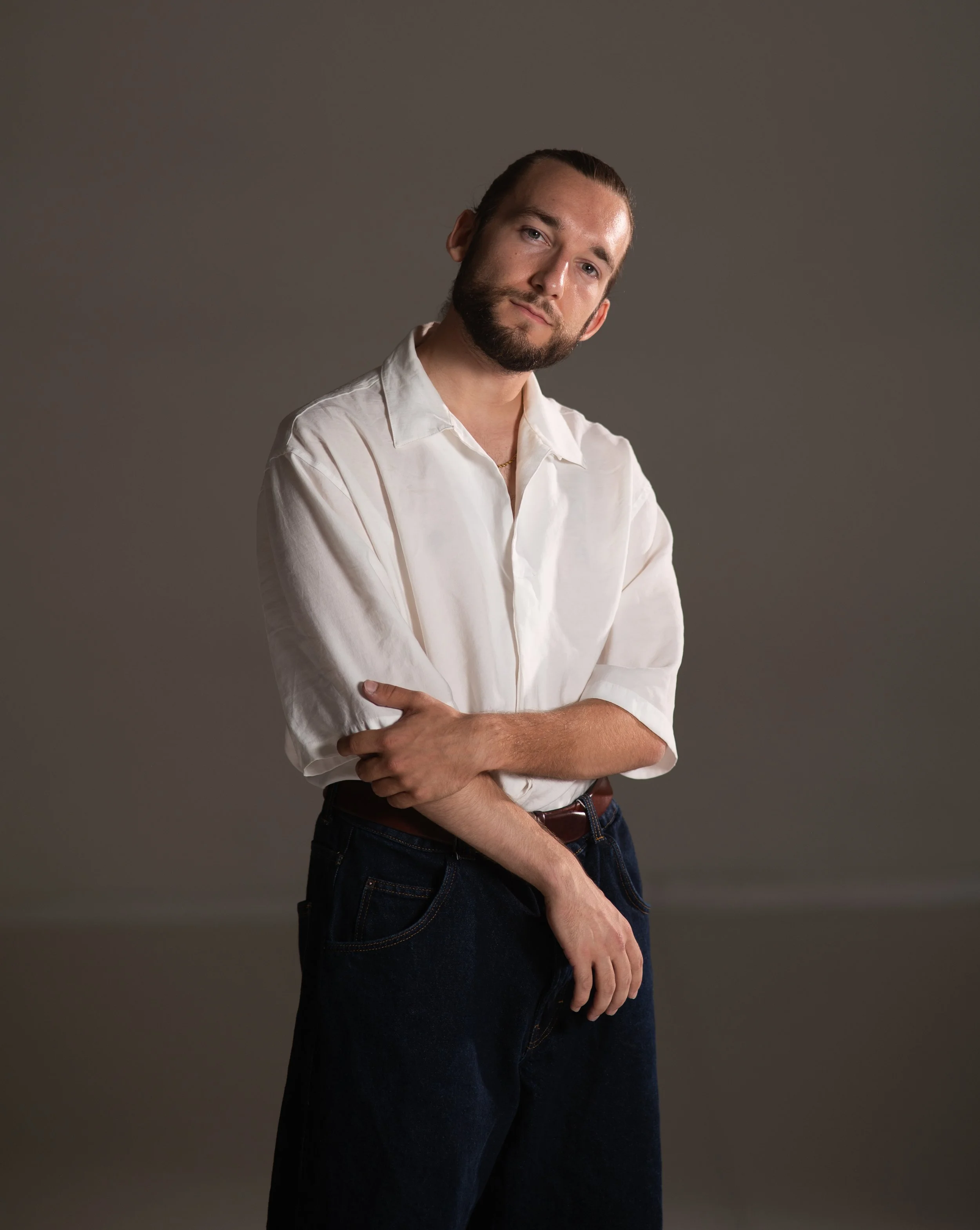 Bearded man in a white shirt and dark jeans posing with crossed arms against a brown background