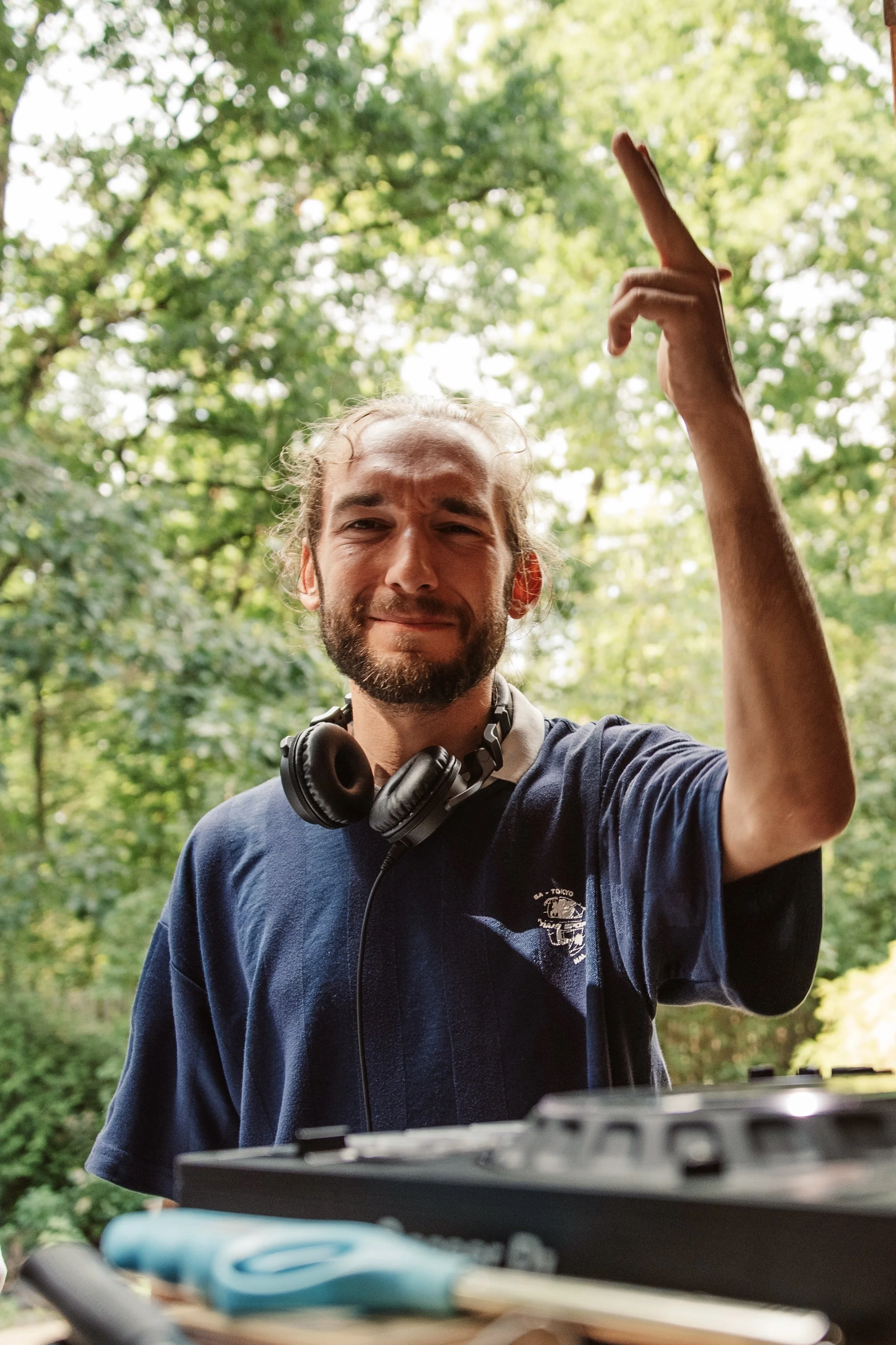 Bearded man wearing headphones raises one finger outdoors in a sunlit forest.