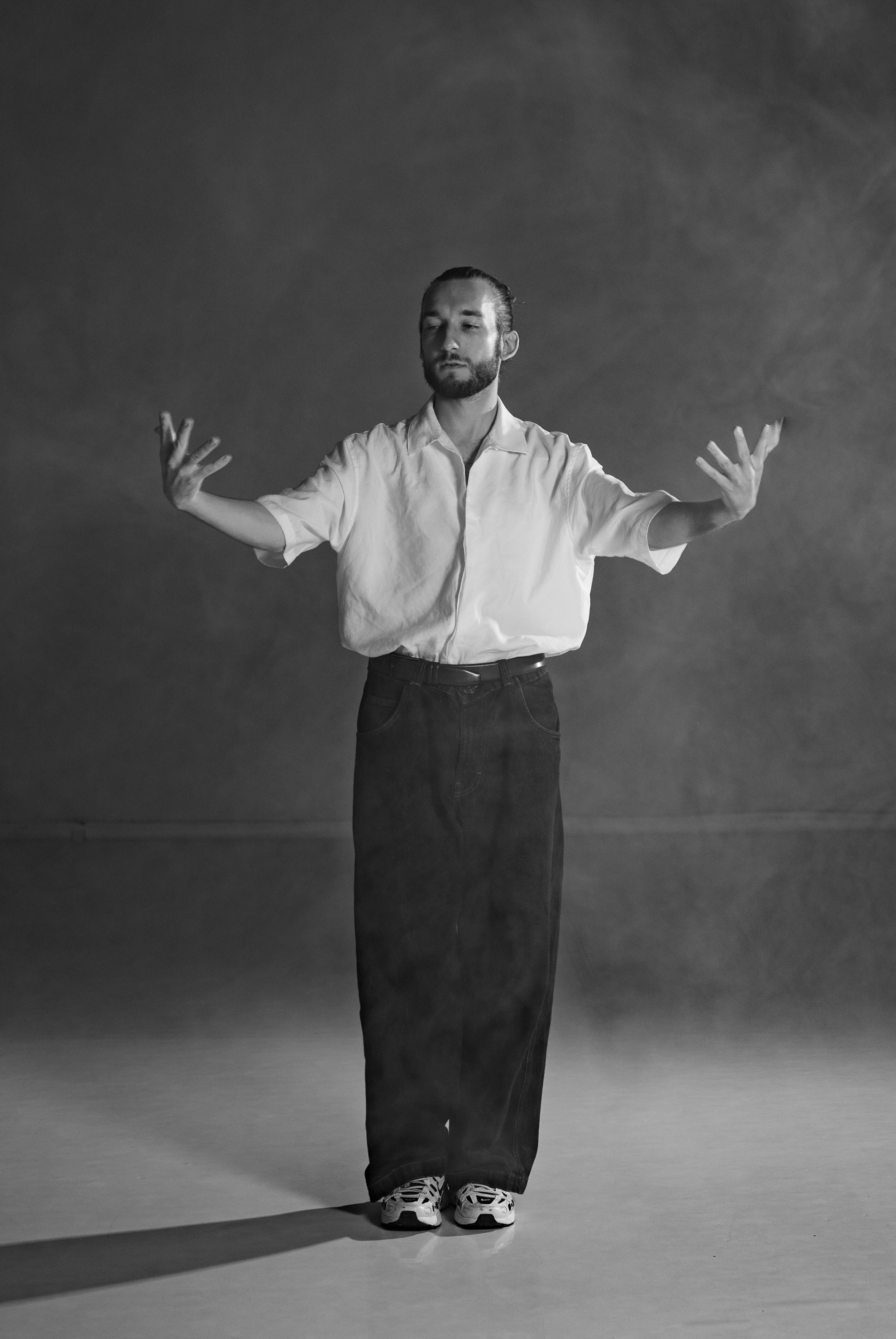 Black-and-white portrait of a bearded man in a white shirt and black pants standing with his arms outstretched against a dark wall.