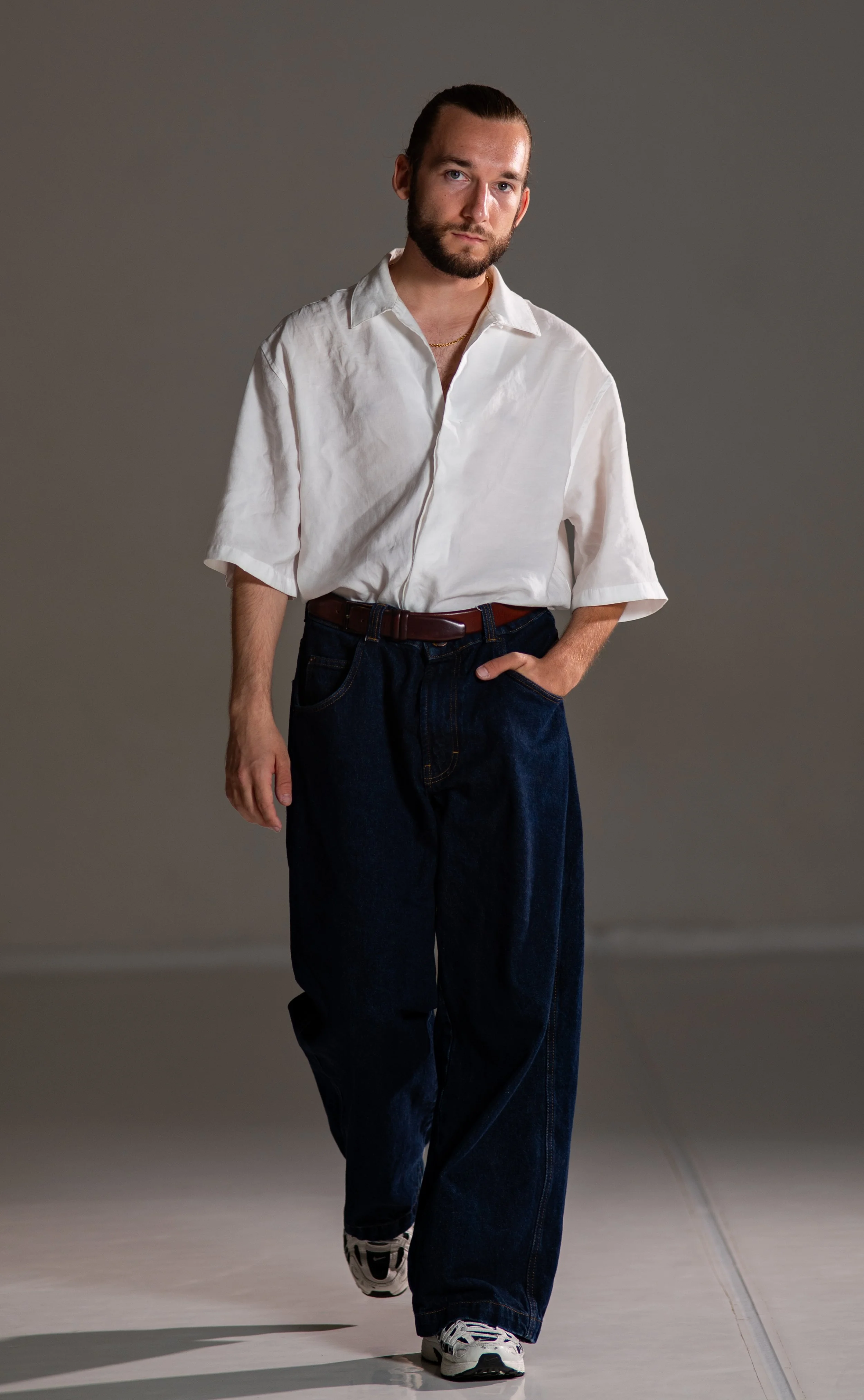 Bearded man in a white shirt, dark jeans, belt, and sneakers standing indoors against a gray wall