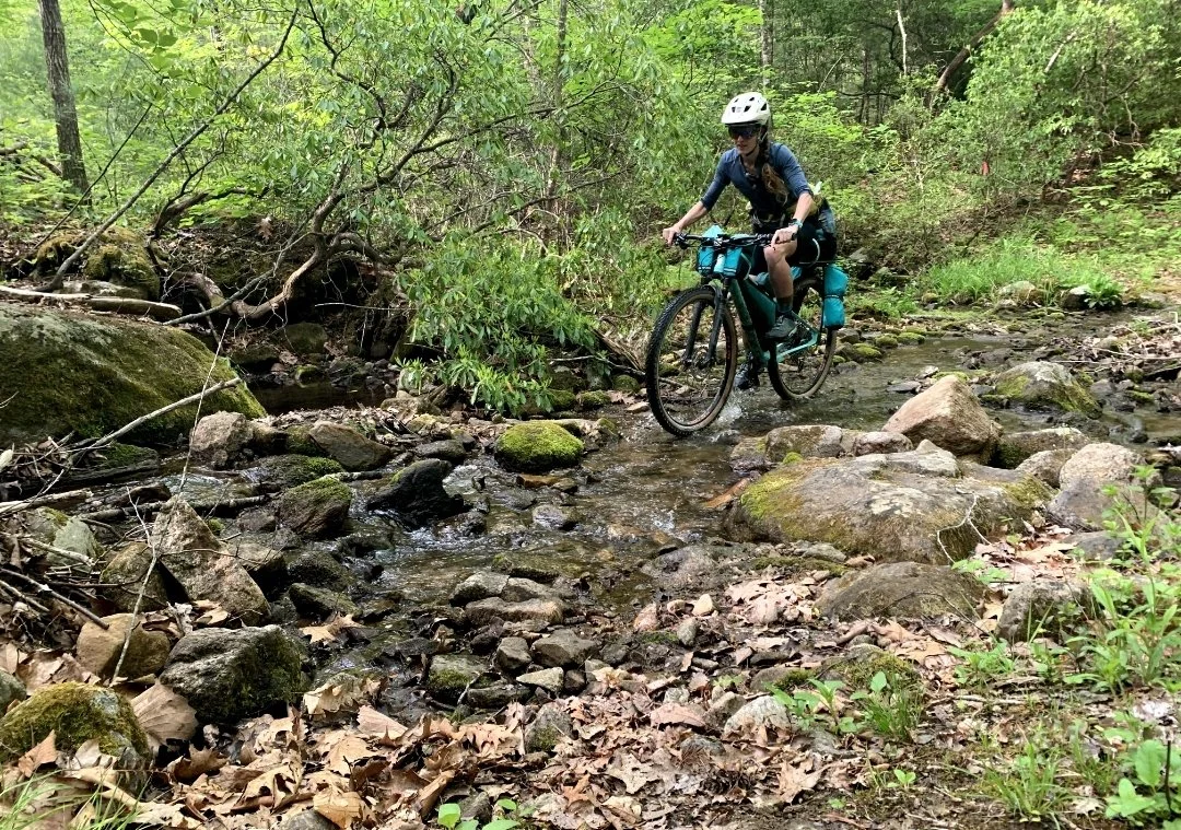 Bikepacker with matching custom bag set rides a loaded bike through a rocky forest creek.