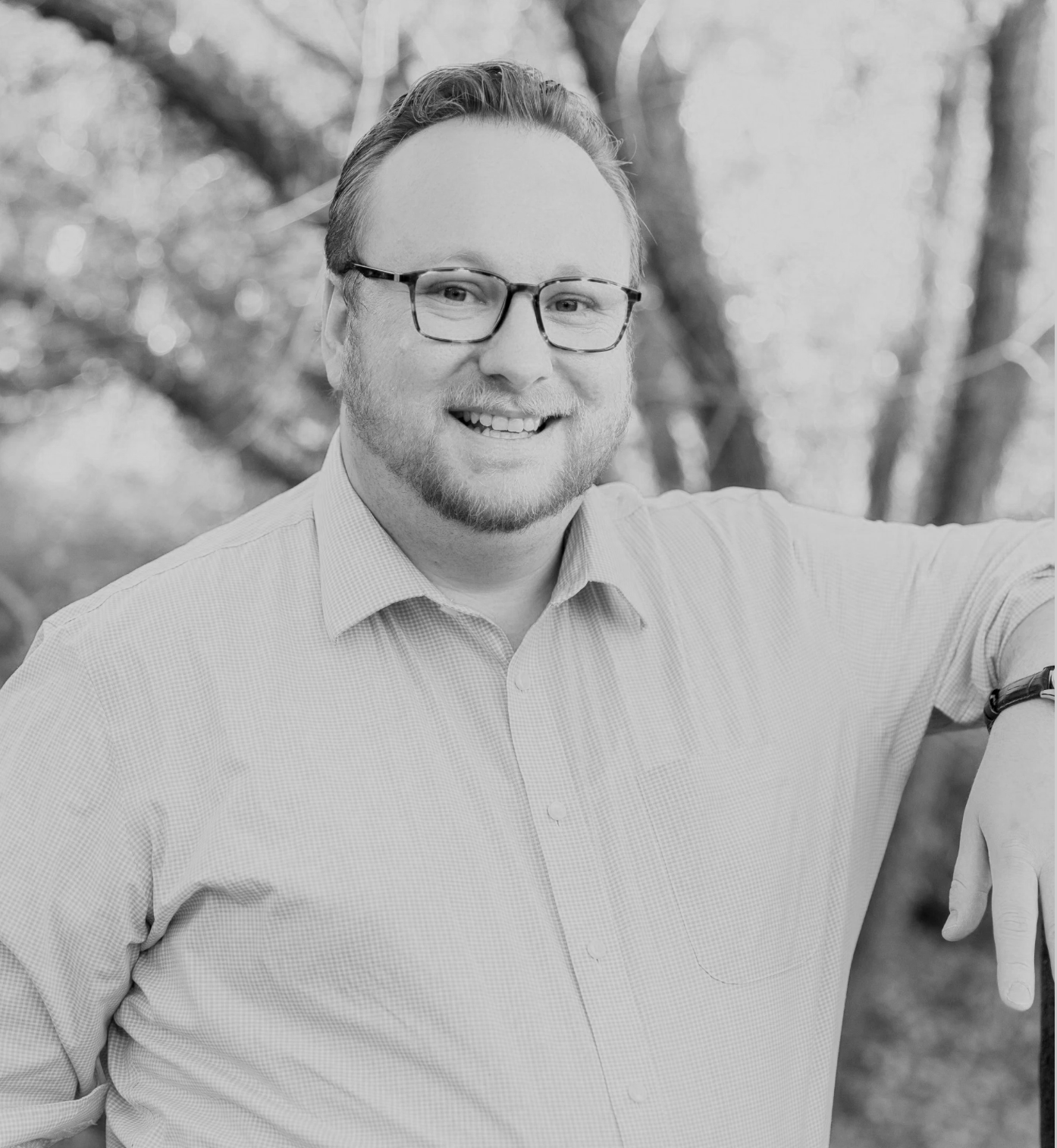 Smiling man wearing glasses and a button-down shirt outdoors in a wooded setting