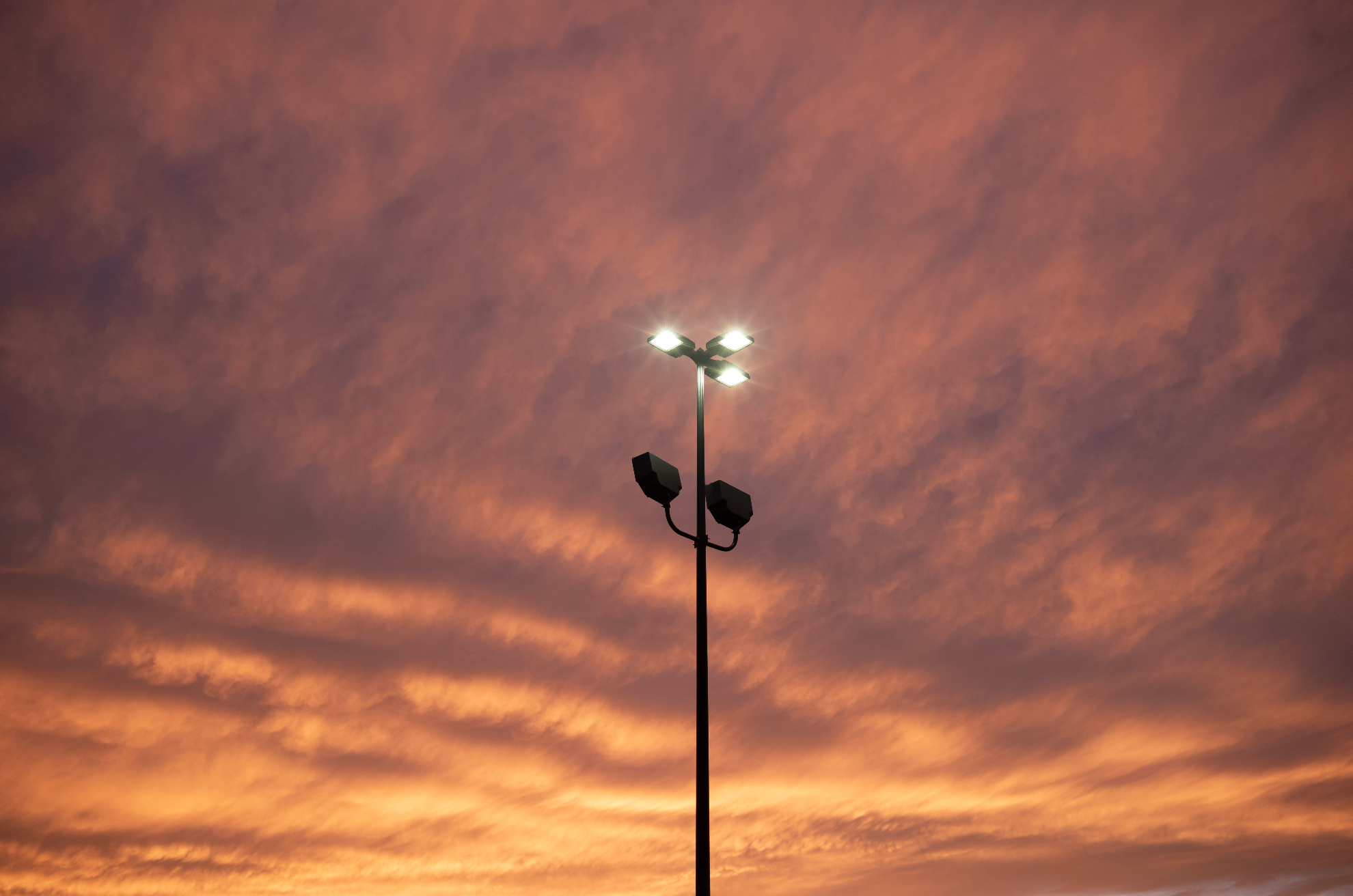 Outdoor stadium light pole illuminated against dramatic orange sunset clouds