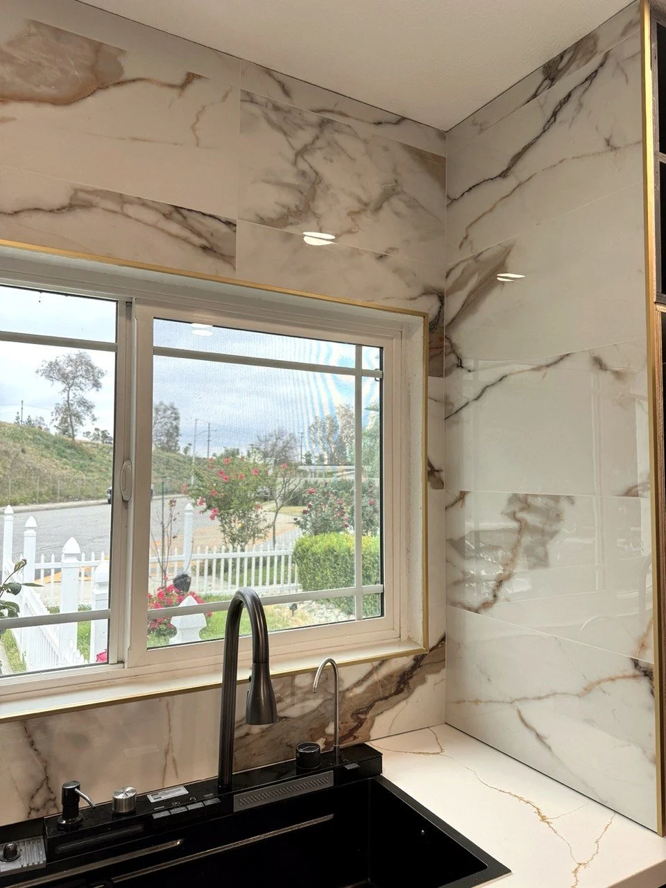 Kitchen sink beneath a window with white marble-look backsplash, black faucet, and outdoor garden view