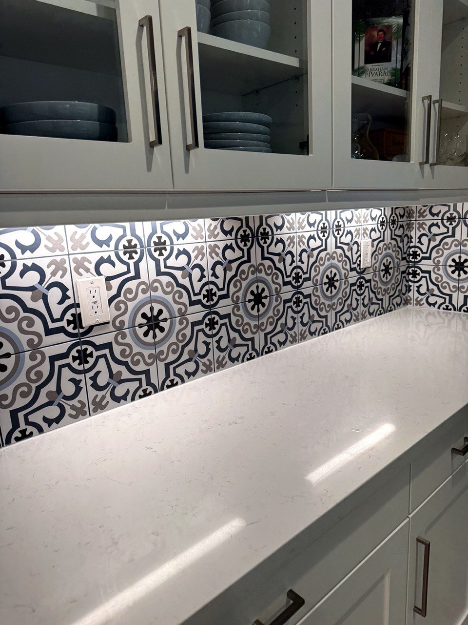White kitchen cabinets with glass doors, stacked dishes, a white countertop, and a blue, gray, and black patterned tile backsplash