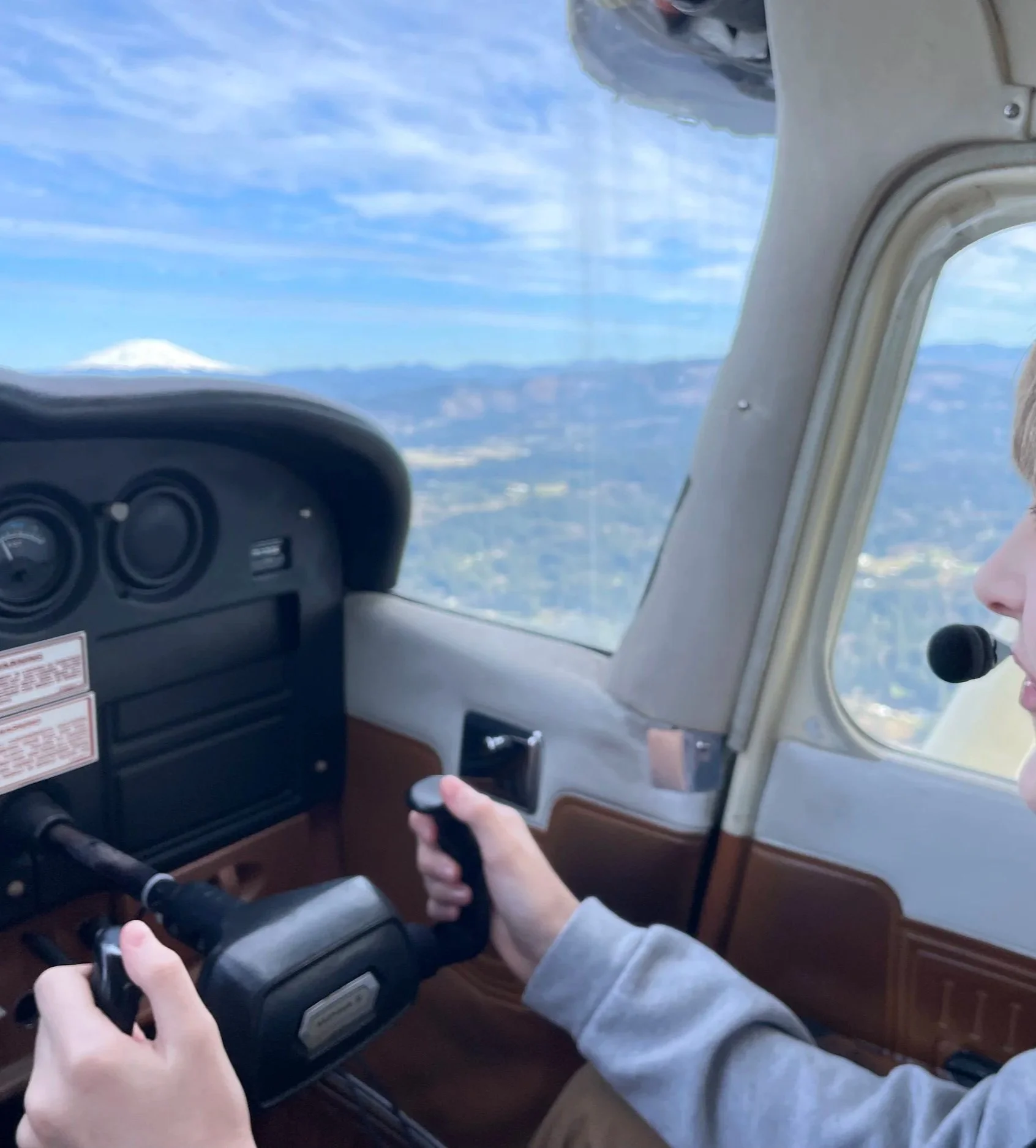 Pilot flying a small airplane over mountainous terrain, with a snow-capped volcano visible through the cockpit window