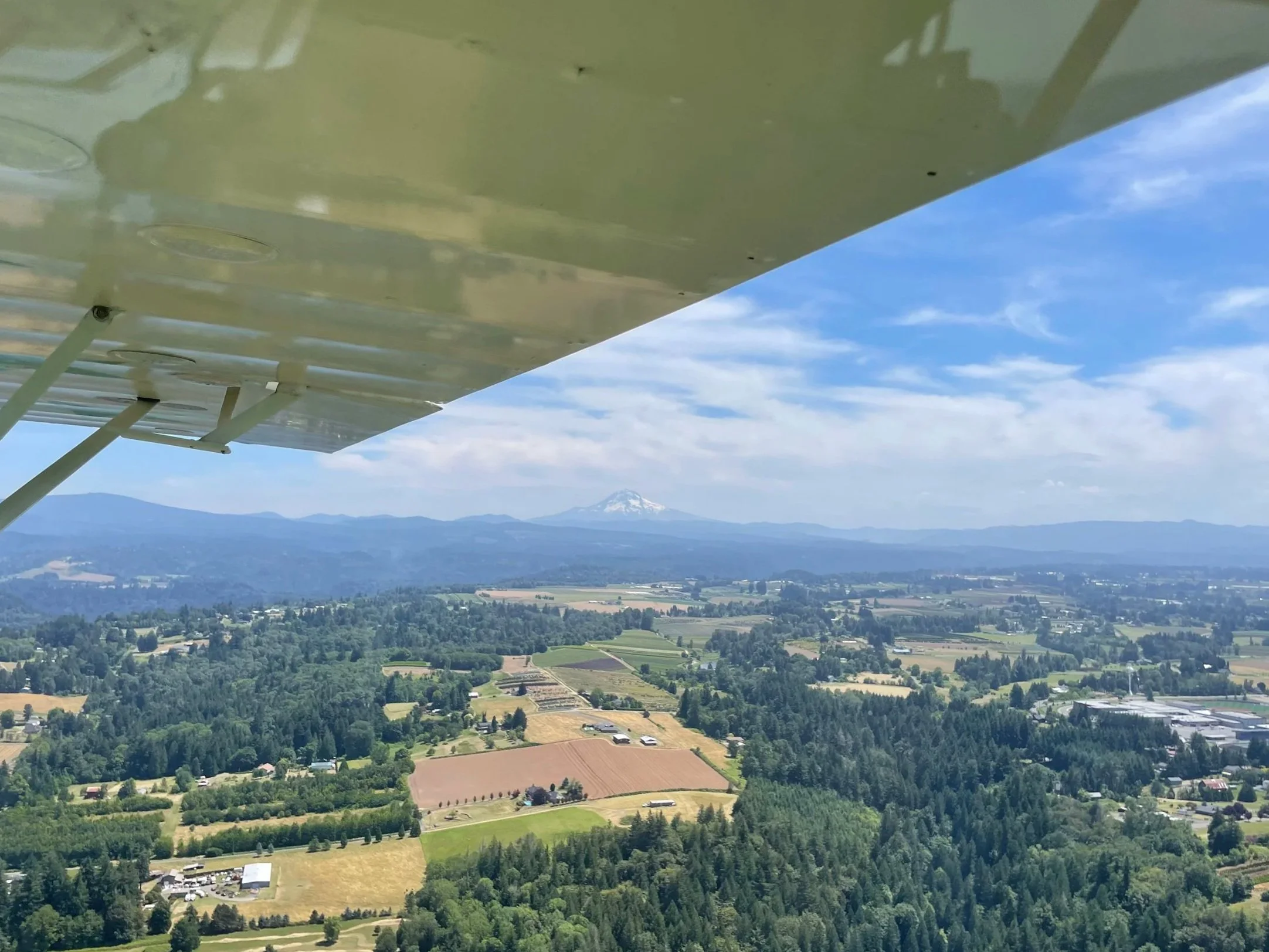 Aerial view from a small aircraft of farmland, forests, and residential areas with a snow-capped volcano in the distance
