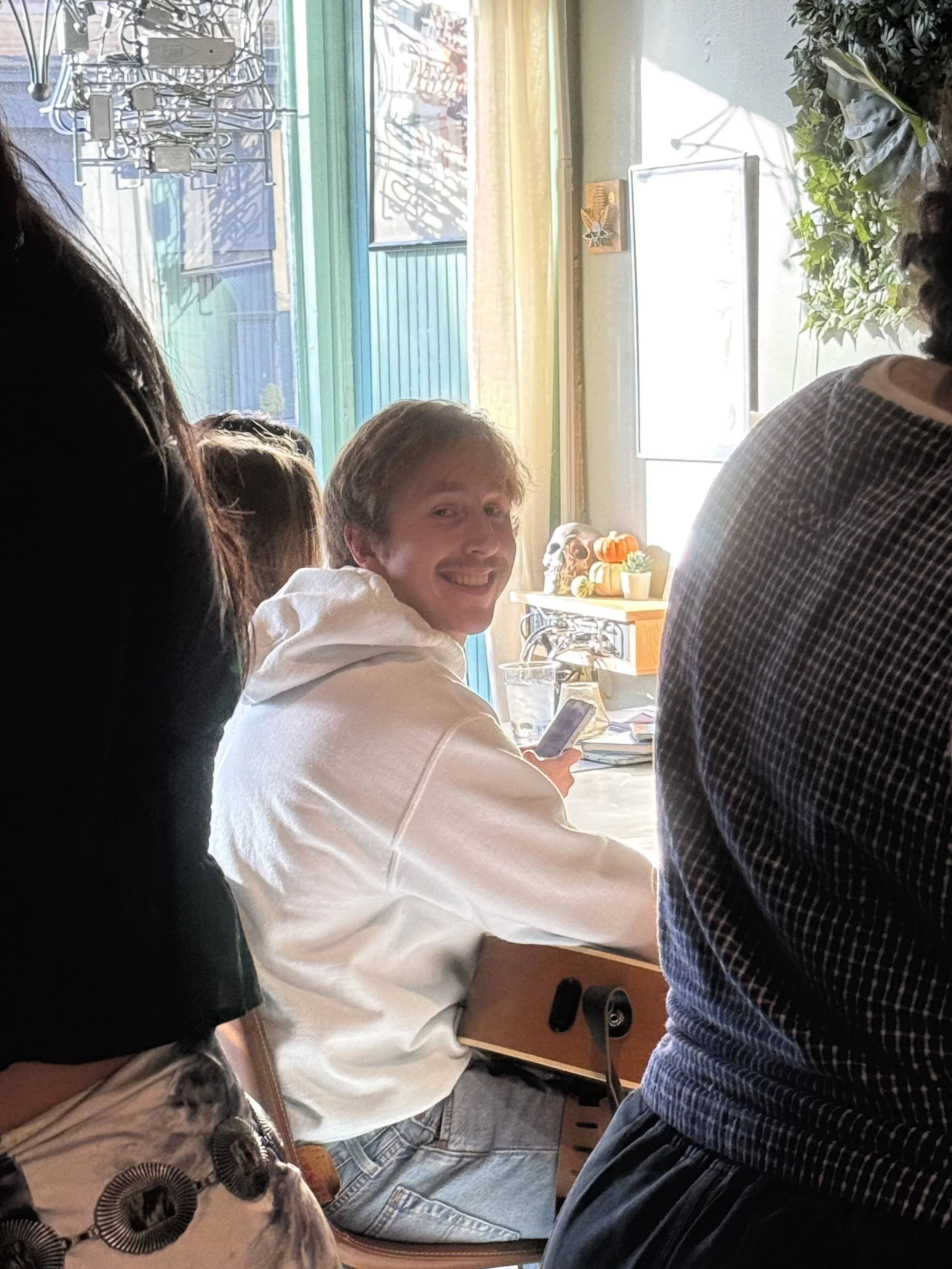 Smiling person in a white hoodie seated indoors with a guitar, surrounded by other people and musical instruments.