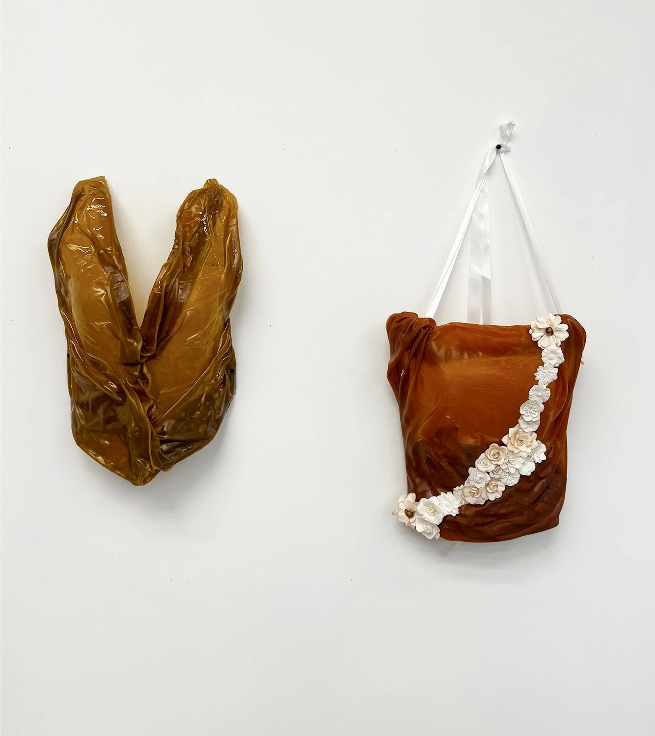 Two amber-brown sculptural wall hangings on a white wall, including a draped piece decorated with a white floral garland.