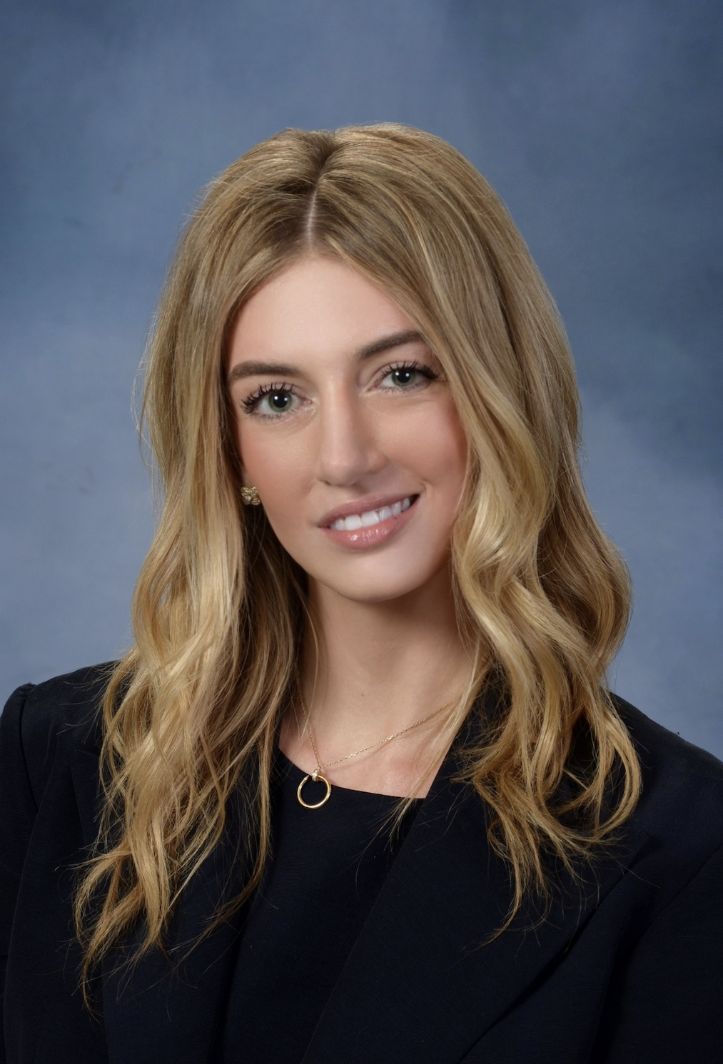 Professional headshot of a smiling blonde woman in a black blazer against a blue-gray background