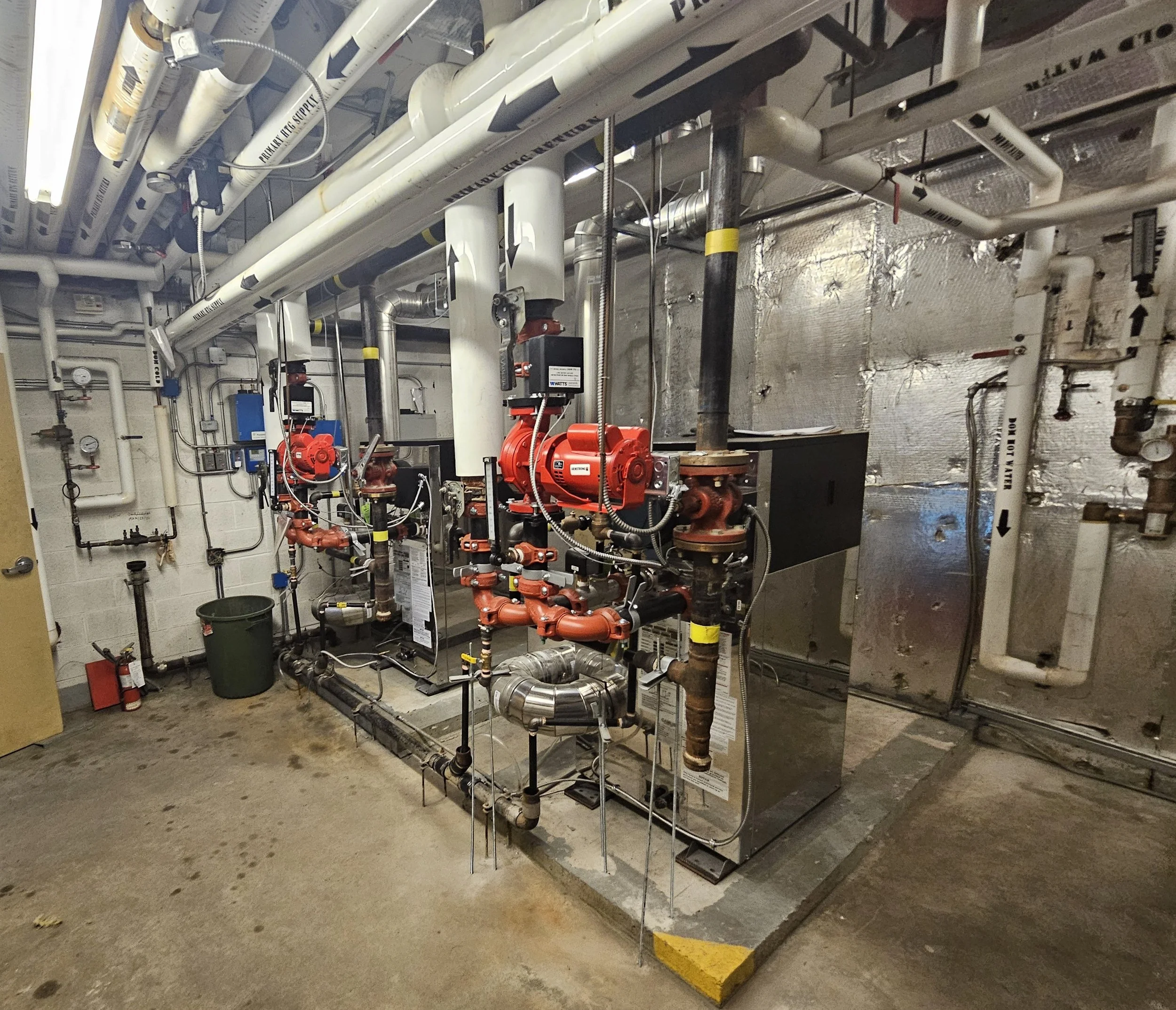 Commercial boiler room with insulated pipes, red circulation pumps, valves, ductwork, and mechanical equipment