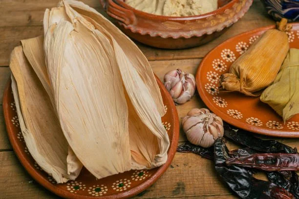 Traditional Mexican ingredients including dried corn husks, garlic, corn, dried chiles, and masa in colorful clay dishes