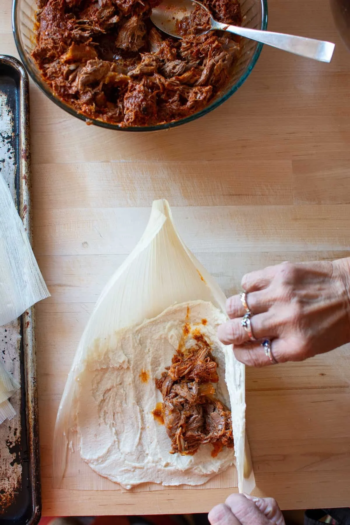 Hands assembling a pulled pork tamale with masa dough and corn husk beside a bowl of shredded meat filling