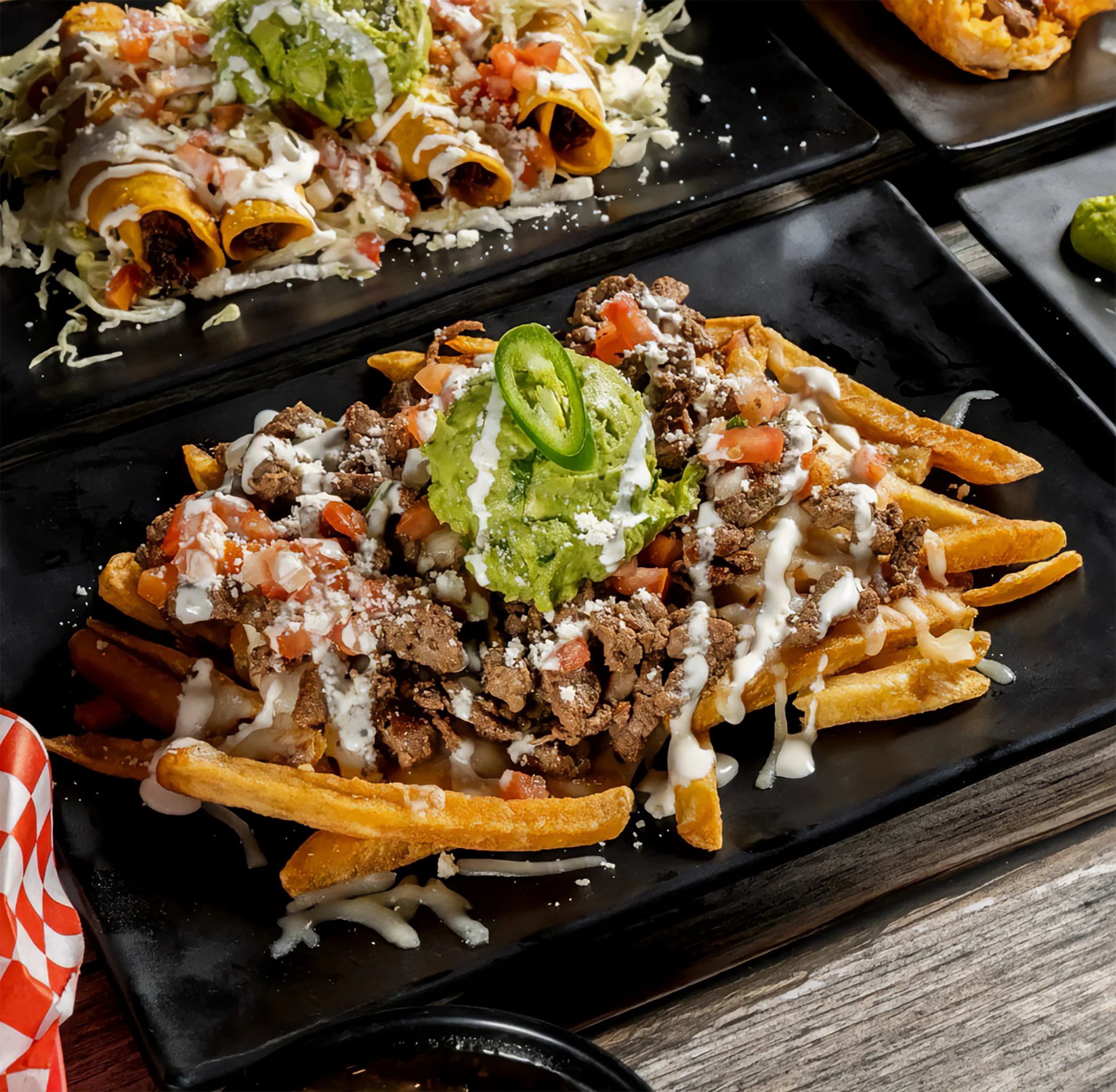 Loaded carne asada fries topped with guacamole, tomatoes, onions, cotija cheese, and creamy sauce, alongside rolled tacos on a black platter