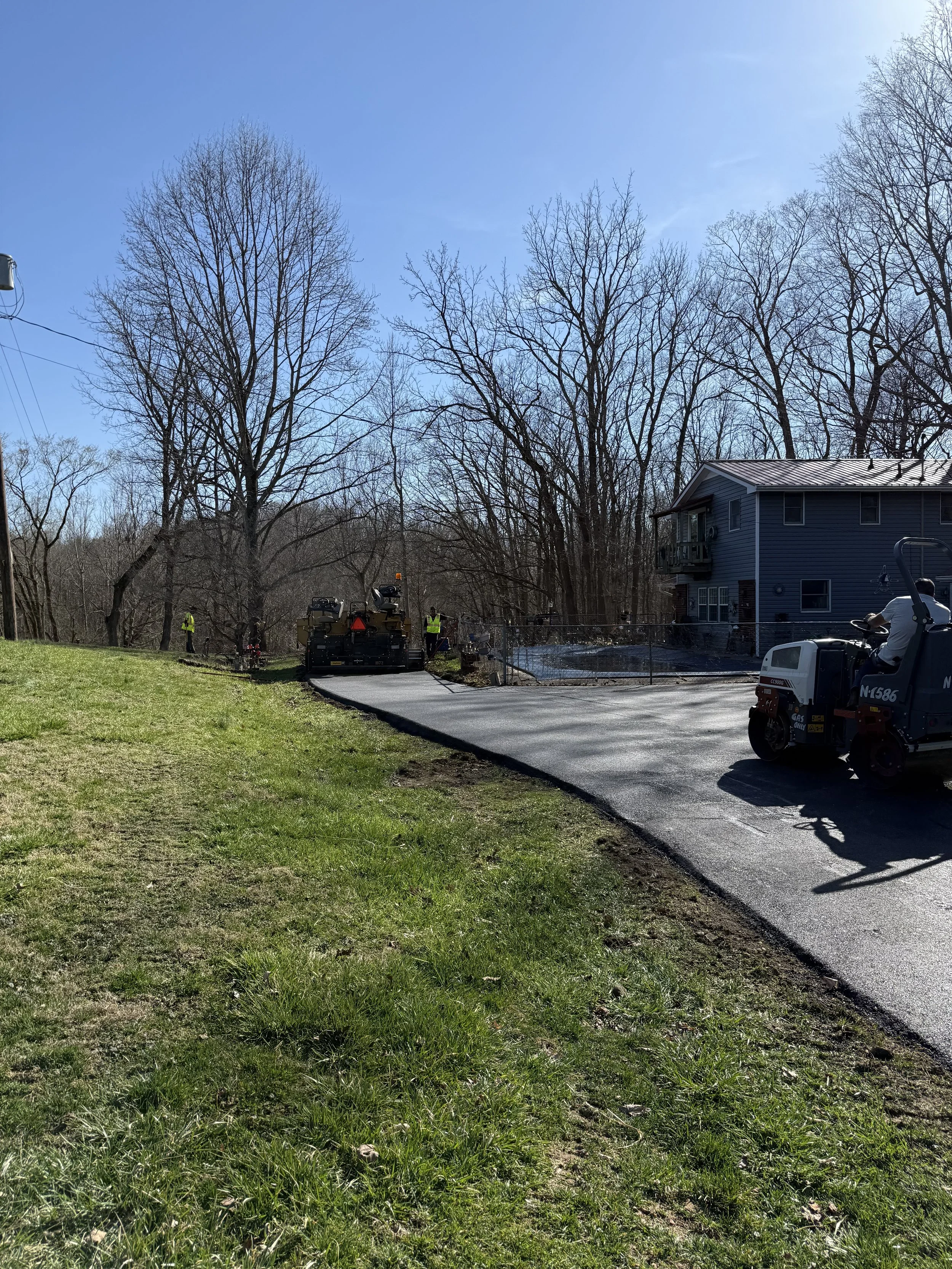 Road construction crew repaves a driveway beside a blue house and leafless trees.