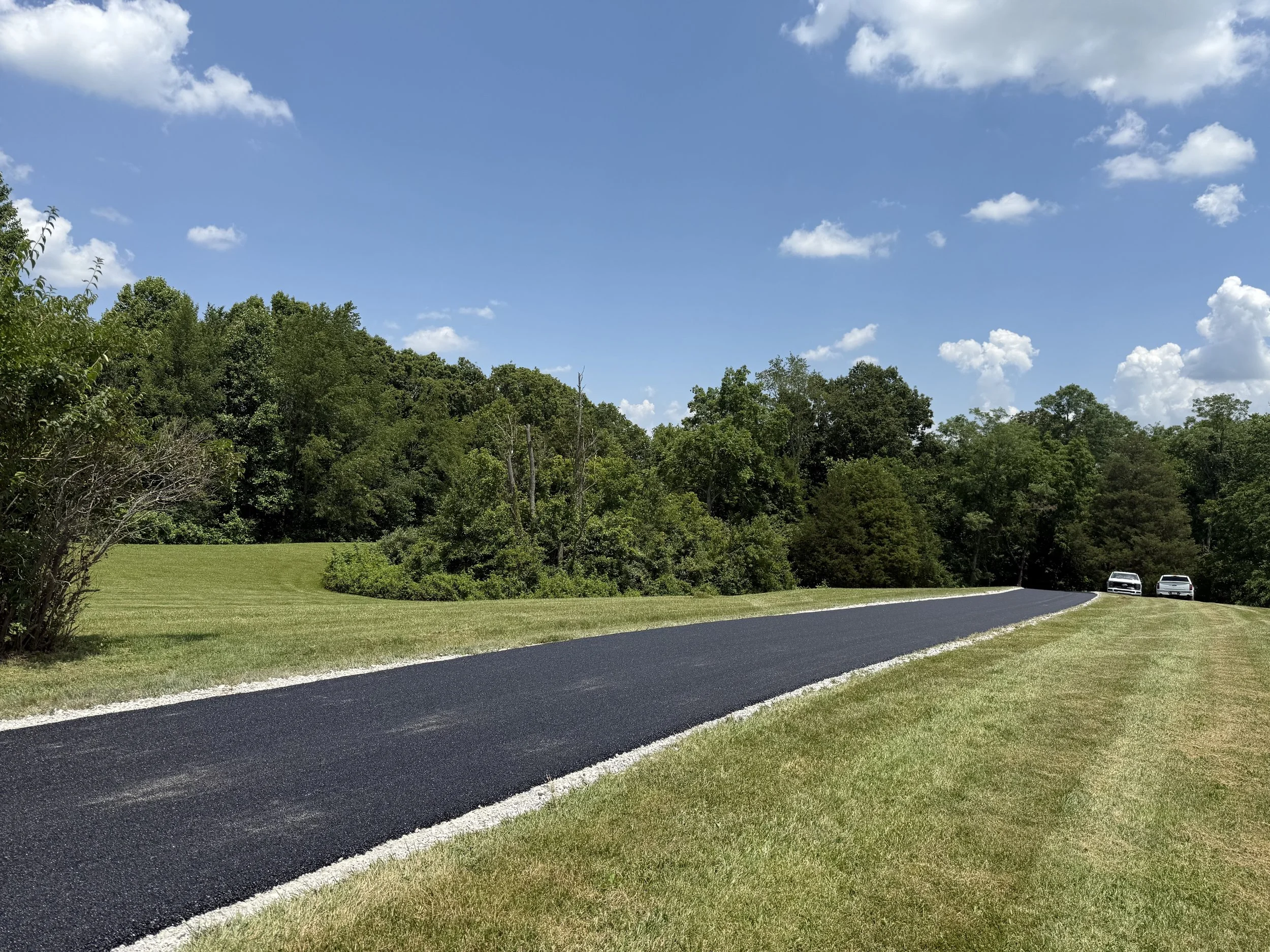 Freshly paved winding driveway through a grassy lawn and wooded landscape under a bright blue sky with clouds