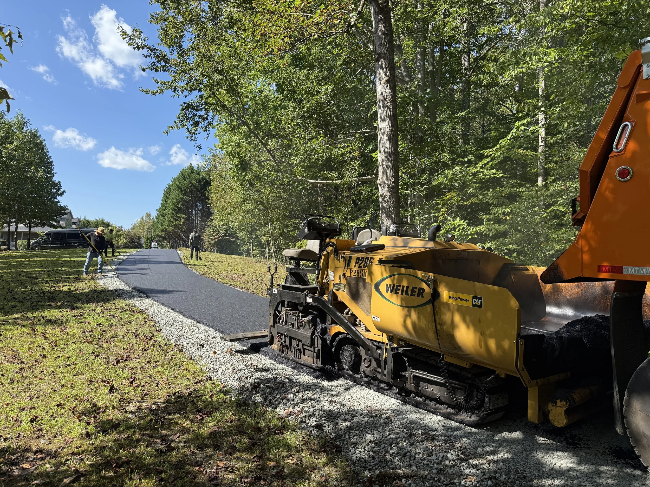 Road paving machine laying fresh asphalt on a residential driveway beside a wooded neighborhood