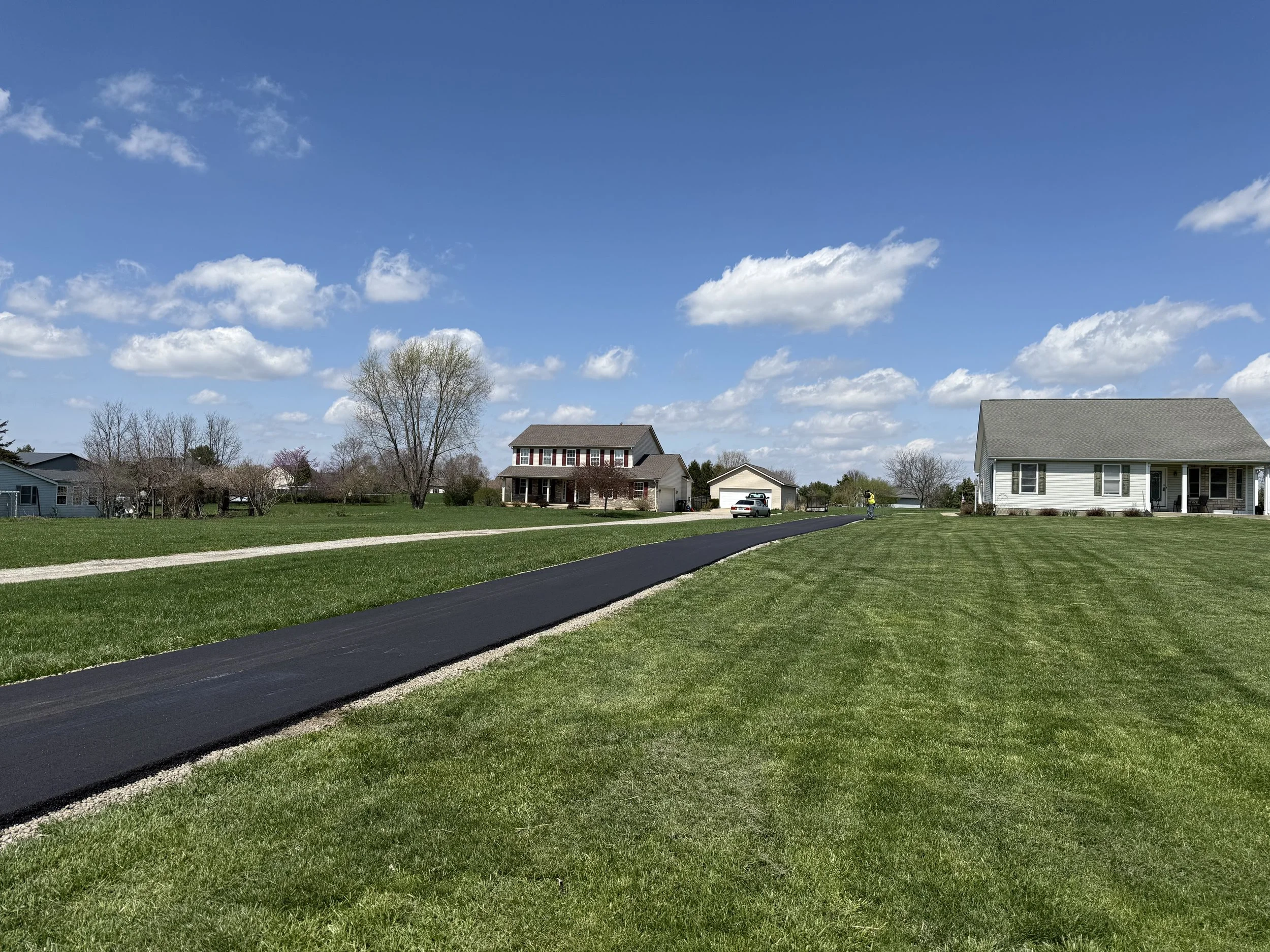 Suburban neighborhood with houses, freshly paved black driveways, green lawns, leafless trees, and a partly cloudy blue sky
