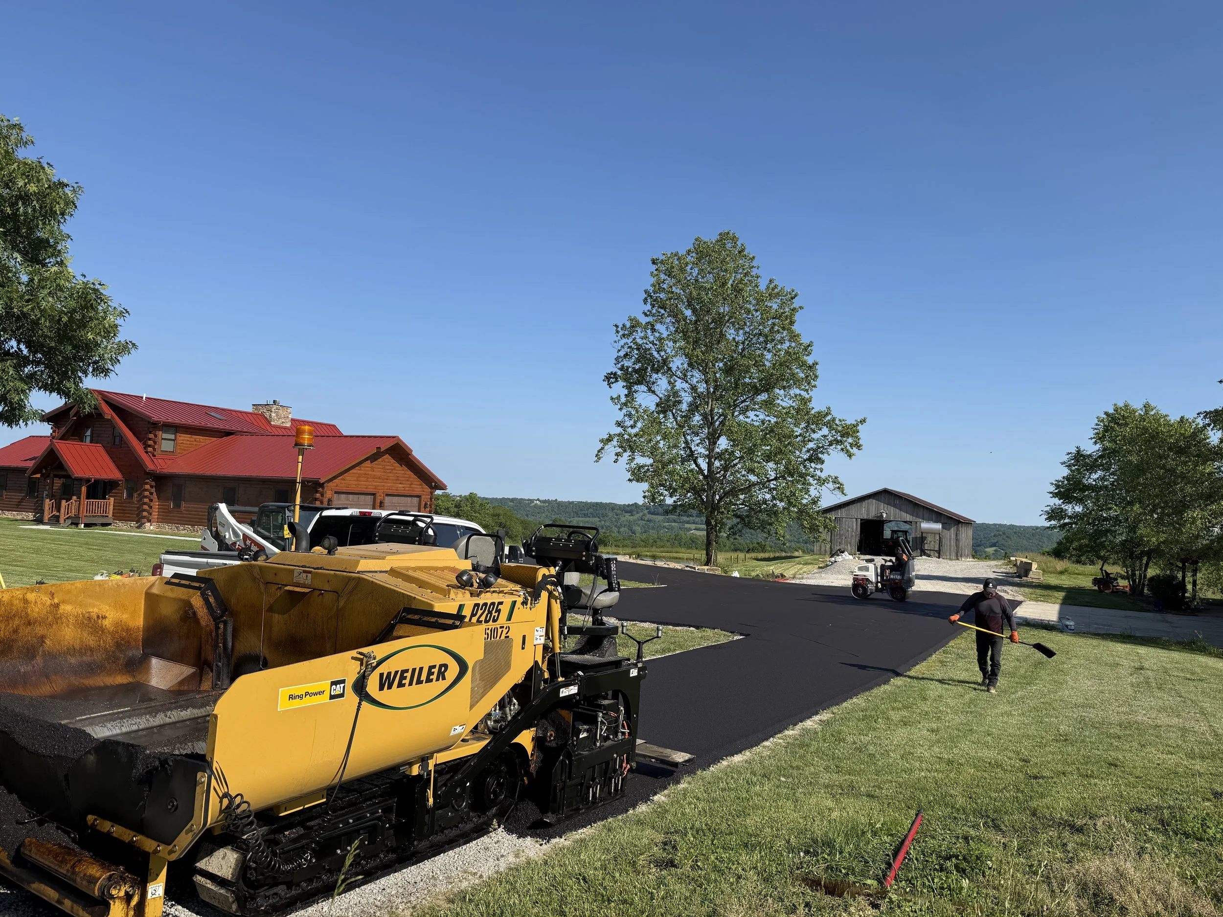 Asphalt paving crew operating a yellow Weiler paver beside a log cabin and barn in a rural landscape