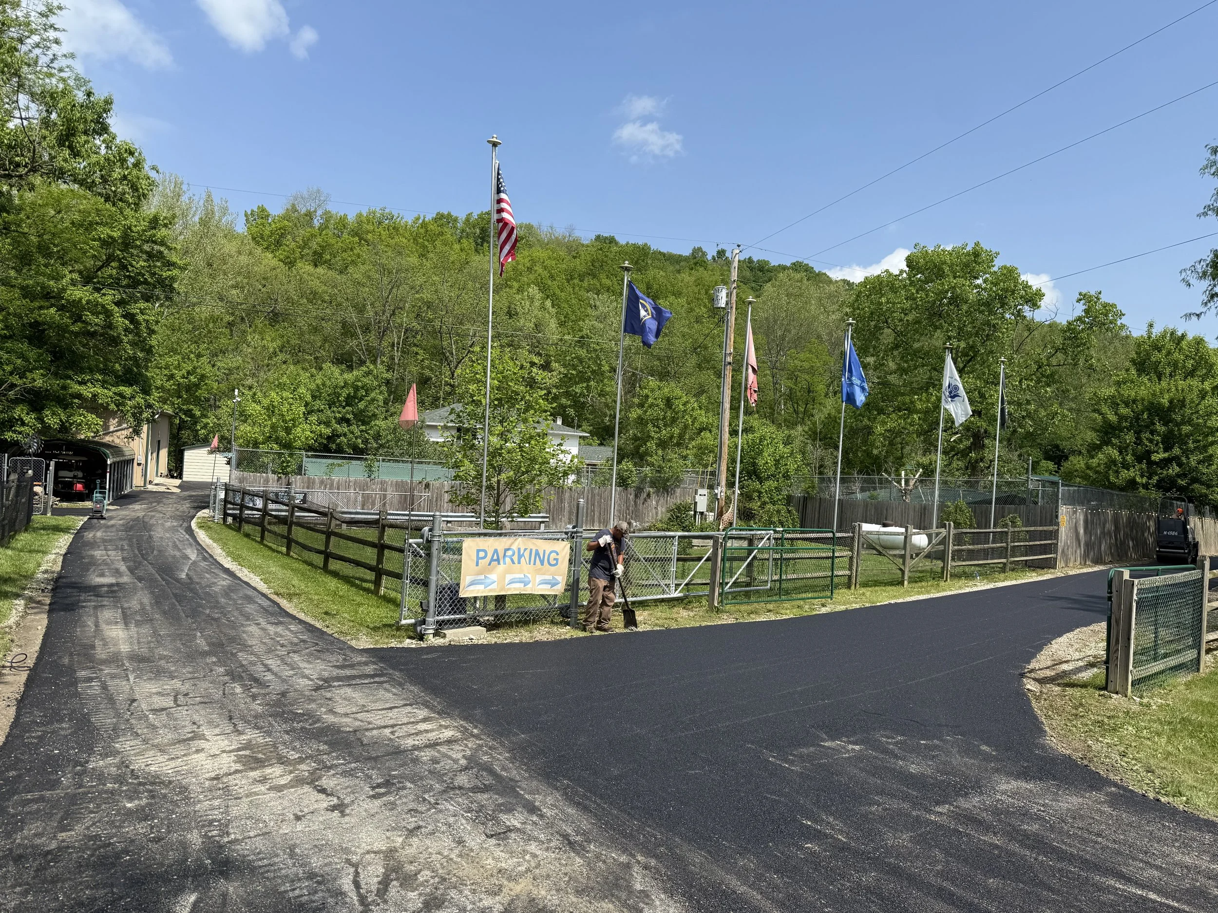 Outdoor facility entrance with American and other flags, fenced grounds, fresh asphalt, and a parking sign on a sunny day