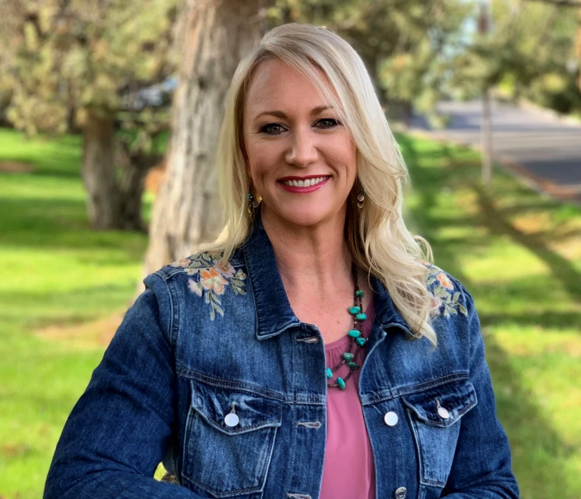 Smiling blonde woman wearing a floral denim jacket and turquoise necklace outdoors in a park