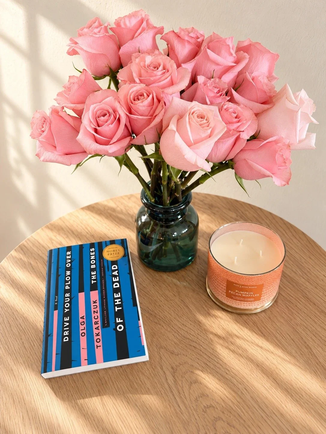 Drive Your Plow Over the Bones of the Dead by Olga Tokarczuk on a wood side table beside pink roses in a teal glass vase and a candle