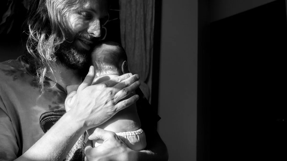 Black-and-white portrait of a bearded father gently holding his newborn baby.