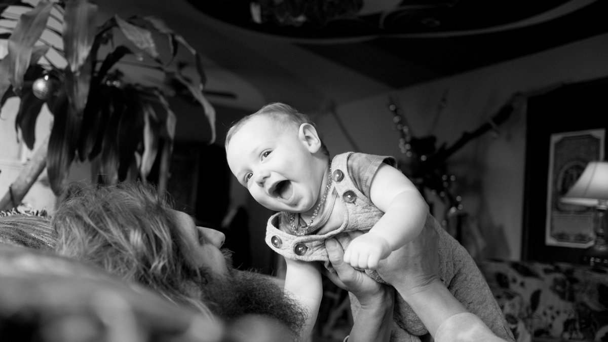 Smiling baby lifted in the air by a bearded adult in a home.