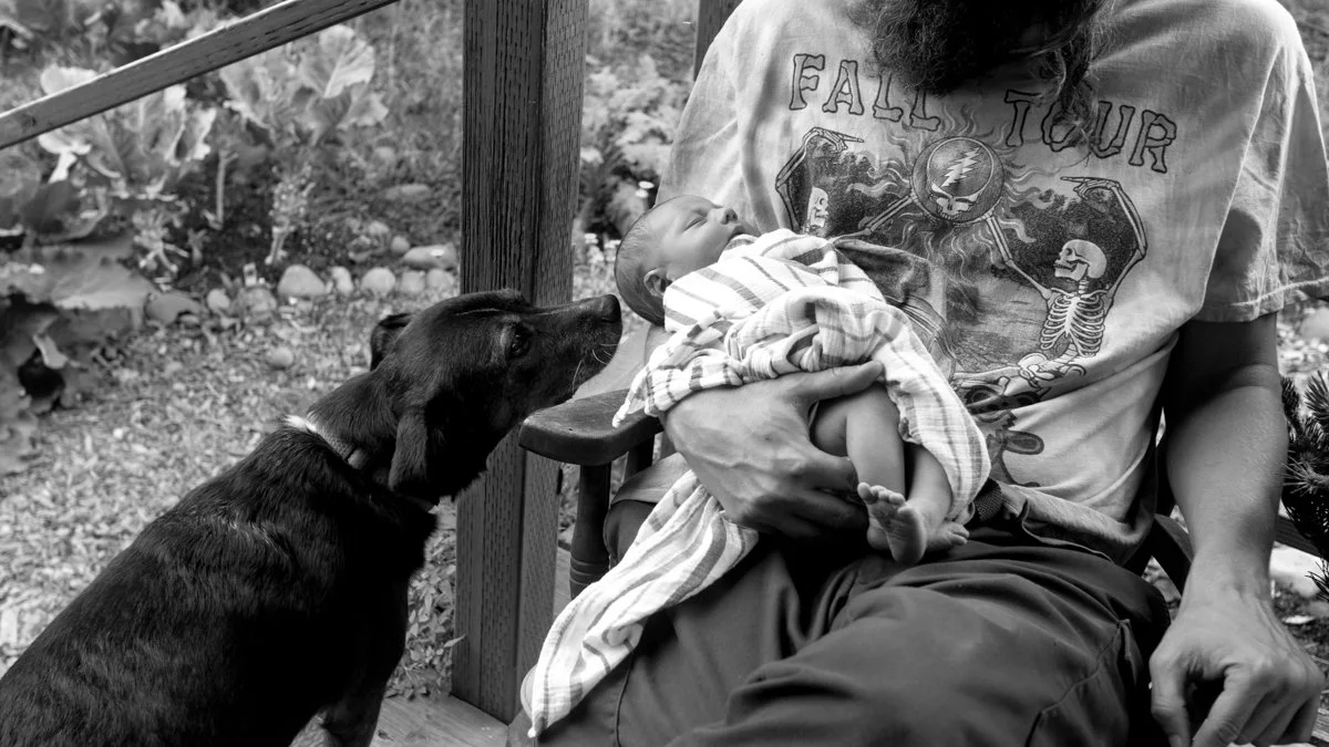 Black-and-white photo of a father holding a newborn baby while a dog looks on from a porch.